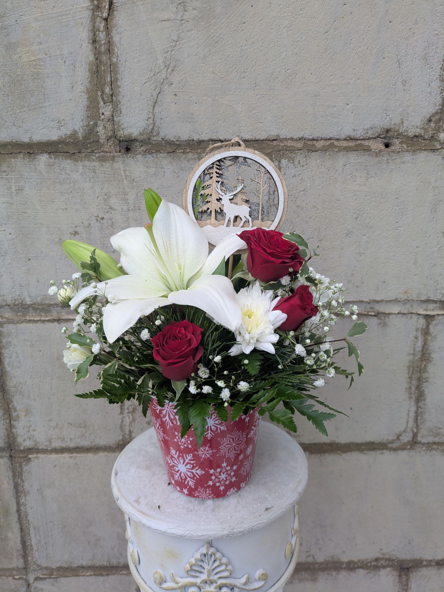 Snowflake Serenade - Bring the peaceful magic of winter indoors with Snowflake Serenade, a graceful Christmas arrangement designed in a charming red-and-white snowflake container. Elegant white lilies take center stage, surrounded by rich red spray roses, fluffy white cushion mums, and delicate clouds of baby’s breath for a soft, fresh, snowy look.  Tucked among the blooms is a festive deer winter-scene pick, adding a touch of woodland wonder that completes the cozy holiday vibe.  Perfect for gifting or brightening your own holiday décor, Snowflake Serenade is a beautiful celebration of winter’s quiet charm.  All flowers, floral colors, and containers are subject to change due to availability.