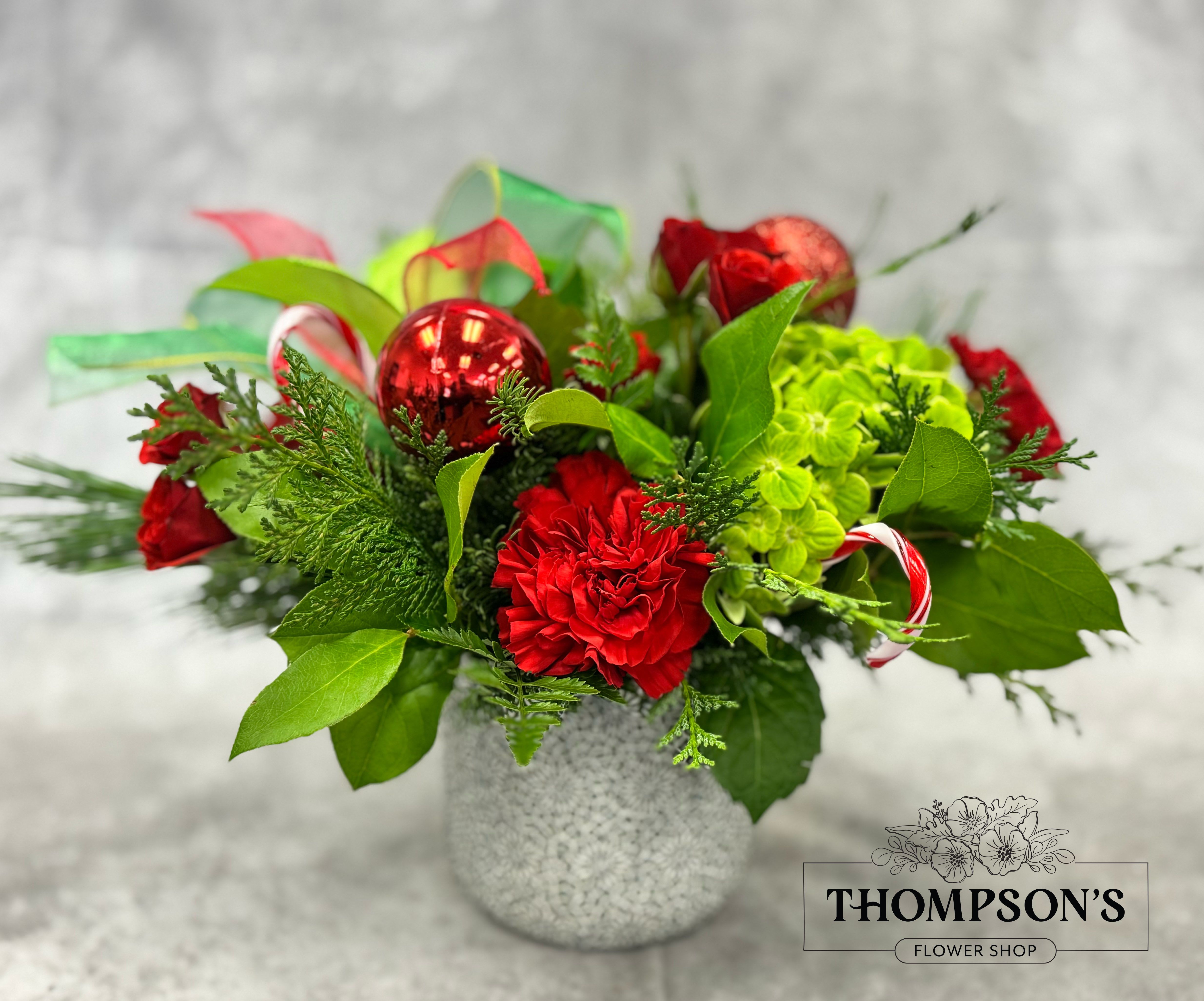 Jolly - Spread some merry cheer with Jolly, a vibrant holiday arrangement bursting with festive color. Red spray roses, carnations, and bright green hydrangea mingle with winter greens, candy canes, and sparkling ornaments for a fun, spirited Christmas design. Finished with a bold red-and-green ribbon, this compact arrangement delivers the joyful energy of the season in every detail. Perfect for gifting, decorating small tables, office desks, kitchen counters, or bringing a playful pop of holiday color to any space.