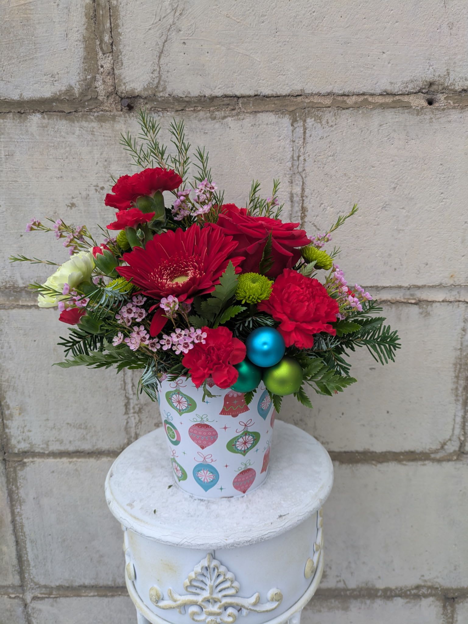 Jolly Jingle - Add a playful pop of Christmas cheer with Jolly Jingle, a fun and festive arrangement tucked inside a vintage ornament–themed container. Bursting with personality, this design mixes bold red roses, a spirited red gerbera daisy, fluffy red carnations, bright red minis, and lively green carnations for a merry color combo. Green button mums and dainty waxflower add texture, while lime, green, and teal baubles bring all the retro jingle-bell vibes.  Whimsical, colorful, and full of holiday fun, Jolly Jingle is guaranteed to spark smiles and spread instant Christmas joy.  All flowers, floral colors, and containers are subject to change due to availability. 