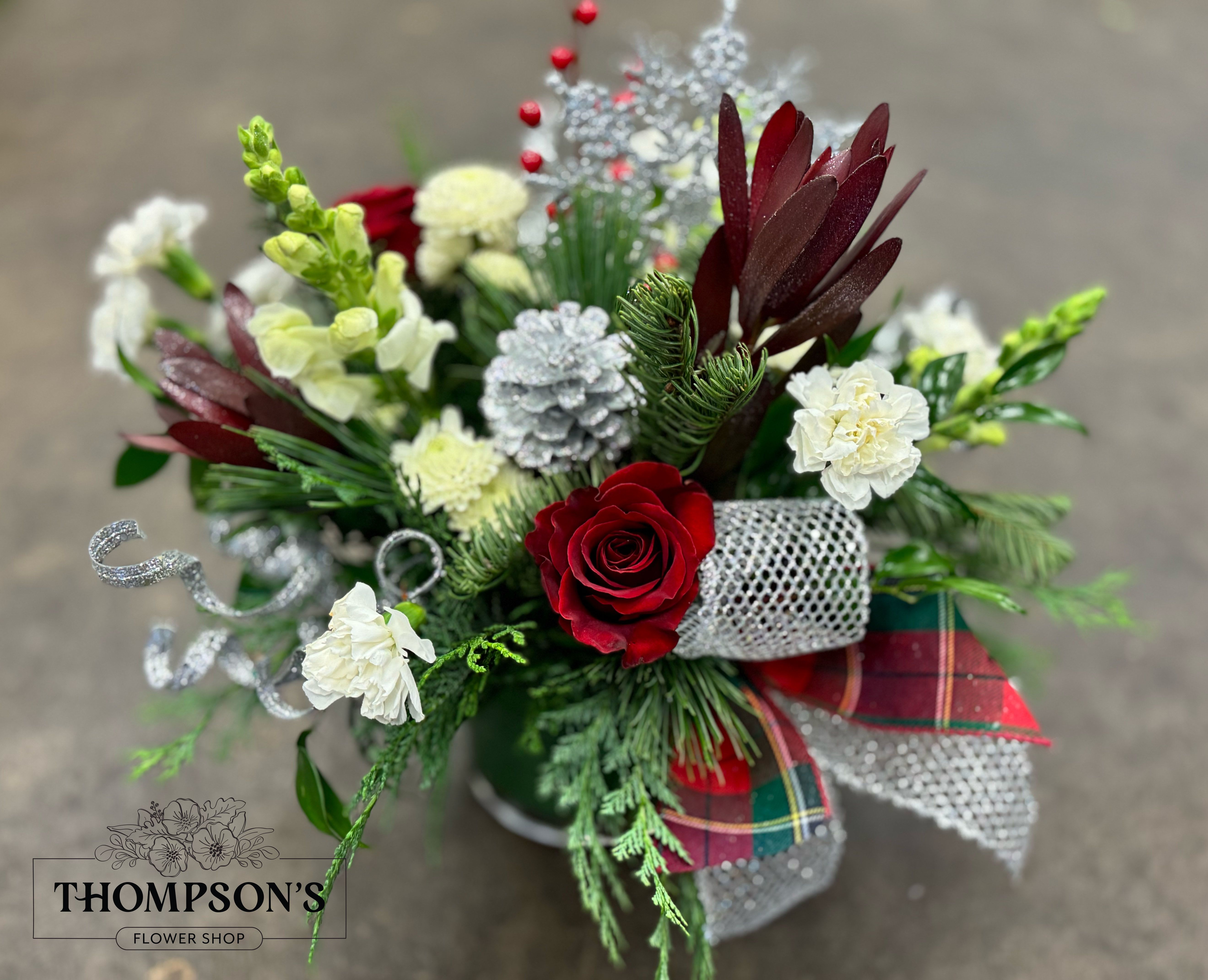 Winter Solstice - Celebrate the serene beauty of the season with Winter Solstice, a radiant holiday arrangement filled with cool shimmer and classic Christmas color. Red roses, white carnations, creamy mums, and winter greens create a lush base, accented with sparkling silver pinecones, glittered curls, snowflake picks, and bright red berries. A touch of plaid ribbon brings warmth and tradition to the design. Perfect for gifting, dining tables, entryways, or bringing a touch of winter magic to any space.