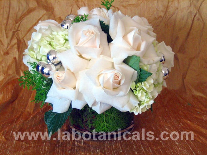 WHITE AS SNOW - Snowy white roses and fluffy hydrangeas are paired with fresh holiday greens and silver ornaments in this charming bouquet. The glass vase is lined with arbor vitae for a pretty touch. Approx. 9" around.