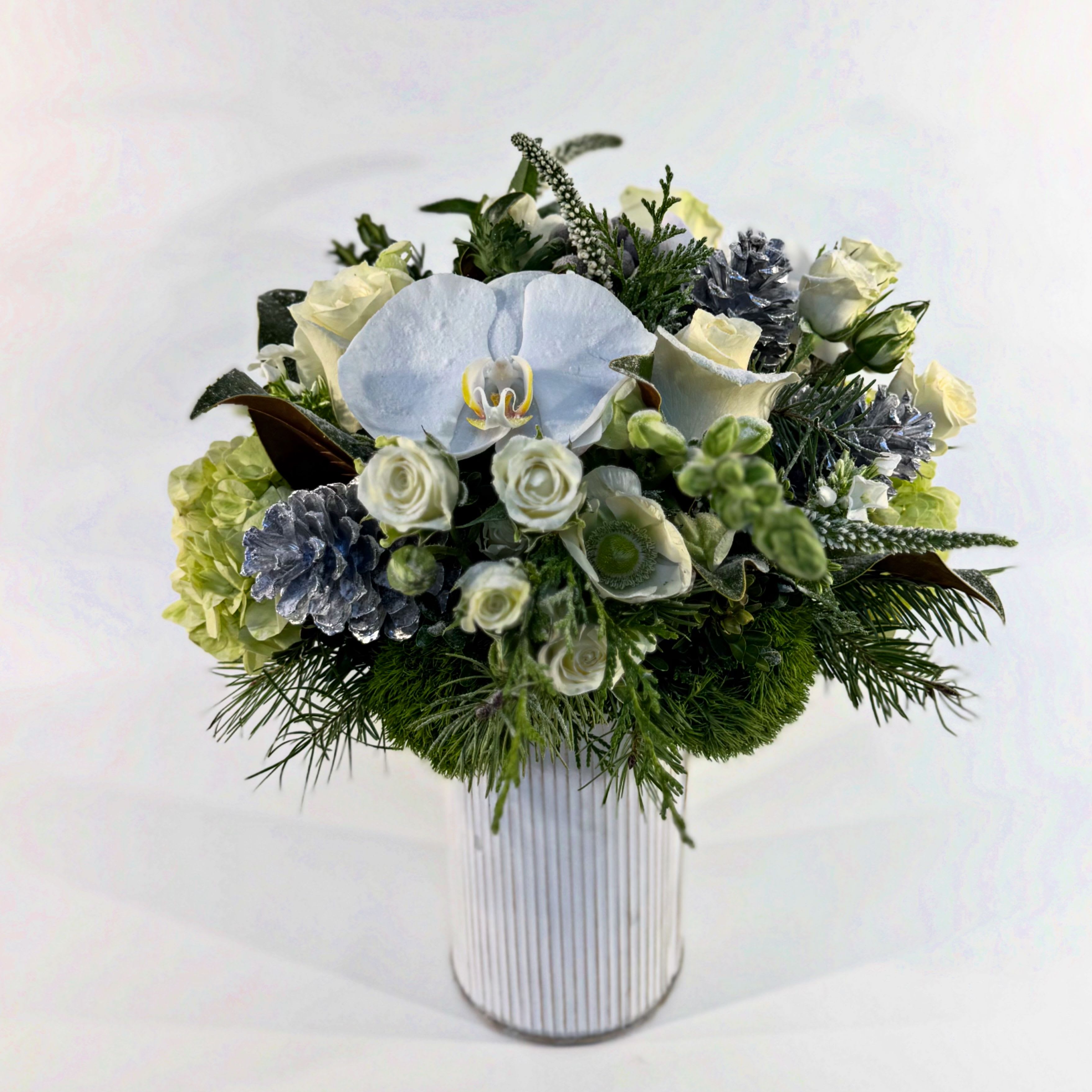 MERRY - A lush winter design incorporating fresh fragrant pine, white and cream blossoms, green hydrangea, silvery pine cones, and velvety magnolia leaves. Designed in a white-washed metal vessel. 