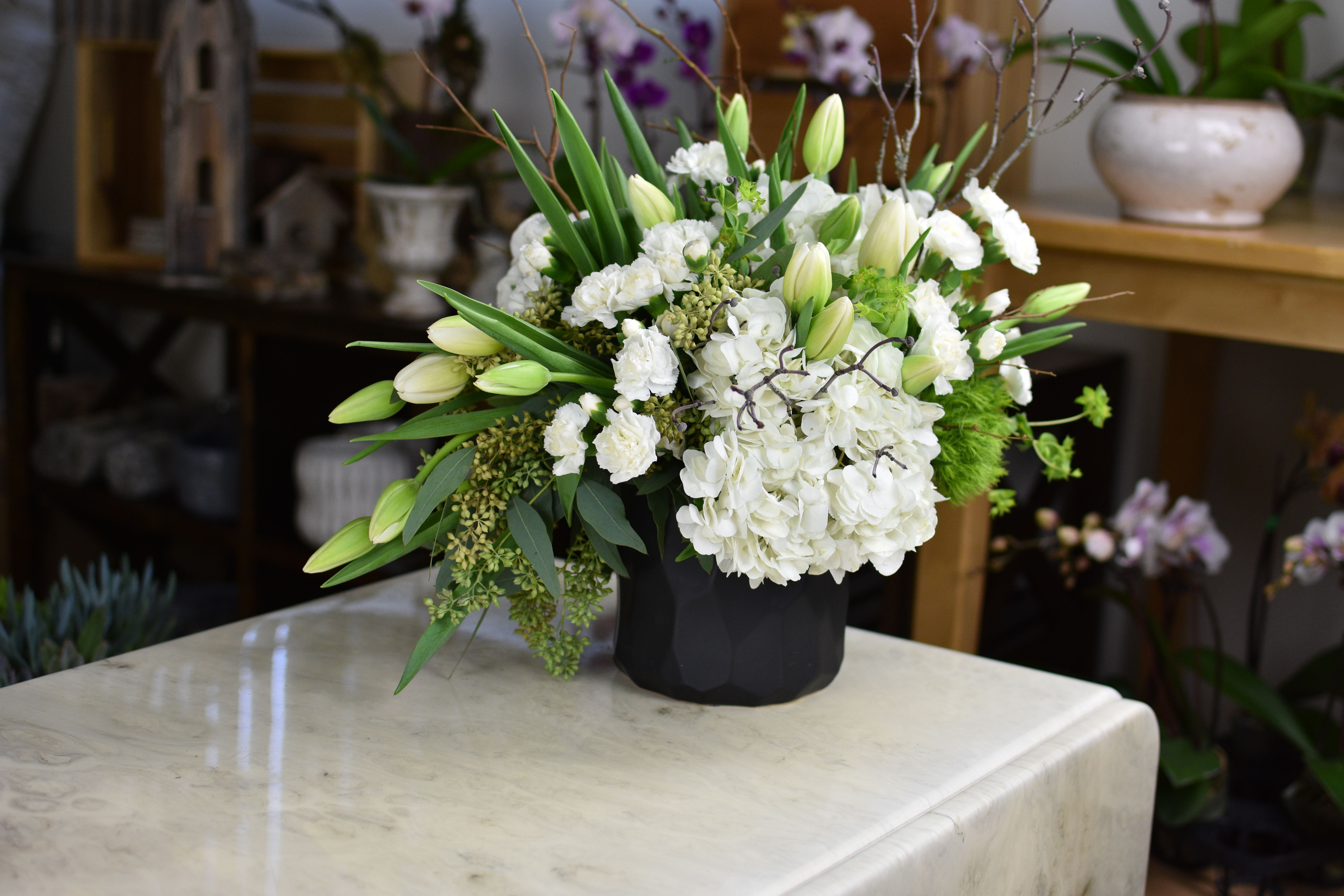 La Maison Blanche - La Maison Blanche Elegant, modern, and beautifully serene, La Maison Blanche is a study in refined white florals arranged with sculptural movement and effortless sophistication. The interplay of soft, rounded blooms and elongated stems creates a balanced, architectural silhouette, while the matte black vessel adds a striking contemporary contrast. This arrangement brings a sense of calm luxury to any space — perfect for sympathy, home décor, or gifting with timeless taste. Please Note: Our arrangements are handcrafted, and no two pieces are ever identical. Variations in flower types, color tones, and exact placement will occur based on daily availability. We do not create factory-duplicate designs — instead, we recreate the style, palette, and overall feel of the arrangement while ensuring every piece is fresh, beautiful, and premium.