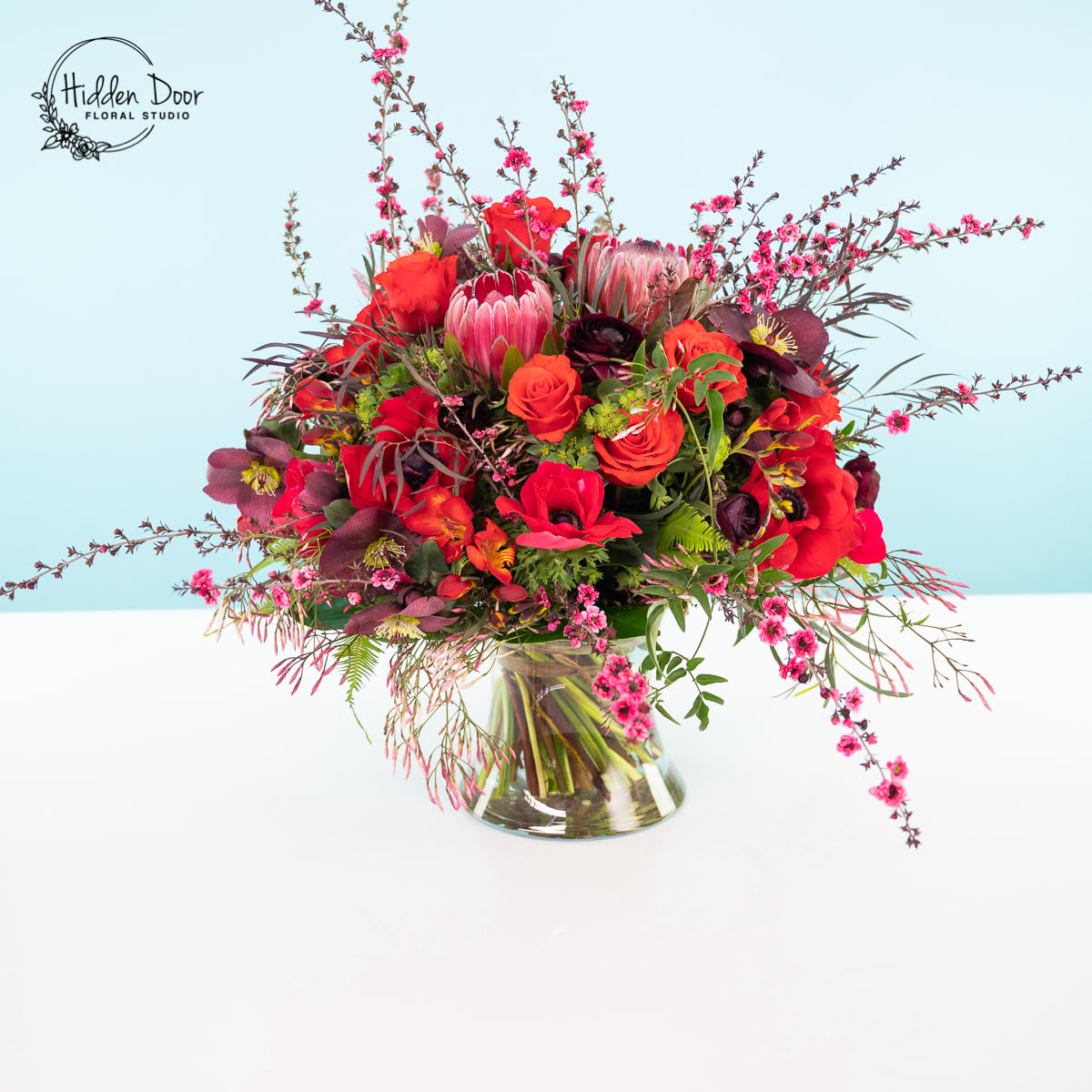 Merlin - Merlin Bold, enchanting, and richly atmospheric, Merlin draws its inspiration from gothic romance and wild, untamed beauty. Deep crimson tones, sculptural textures, and dramatic sweeping lines create a striking, moody composition that feels both magical and sophisticated. Designed in a modern glass vessel, this arrangement evokes the allure of an enchanted forest — powerful, captivating, and full of depth. Perfect for statement gifting, dramatic interiors, or anyone who gravitates toward florals with intensity and artistic flair. Please Note: Our arrangements are handcrafted, and no two pieces are ever identical. Variations in flower types, color tones, and exact placement will occur based on daily availability. We do not create factory-duplicate designs — instead, we recreate the style, palette, and overall feel of the arrangement while ensuring every piece is fresh, beautiful, and premium.