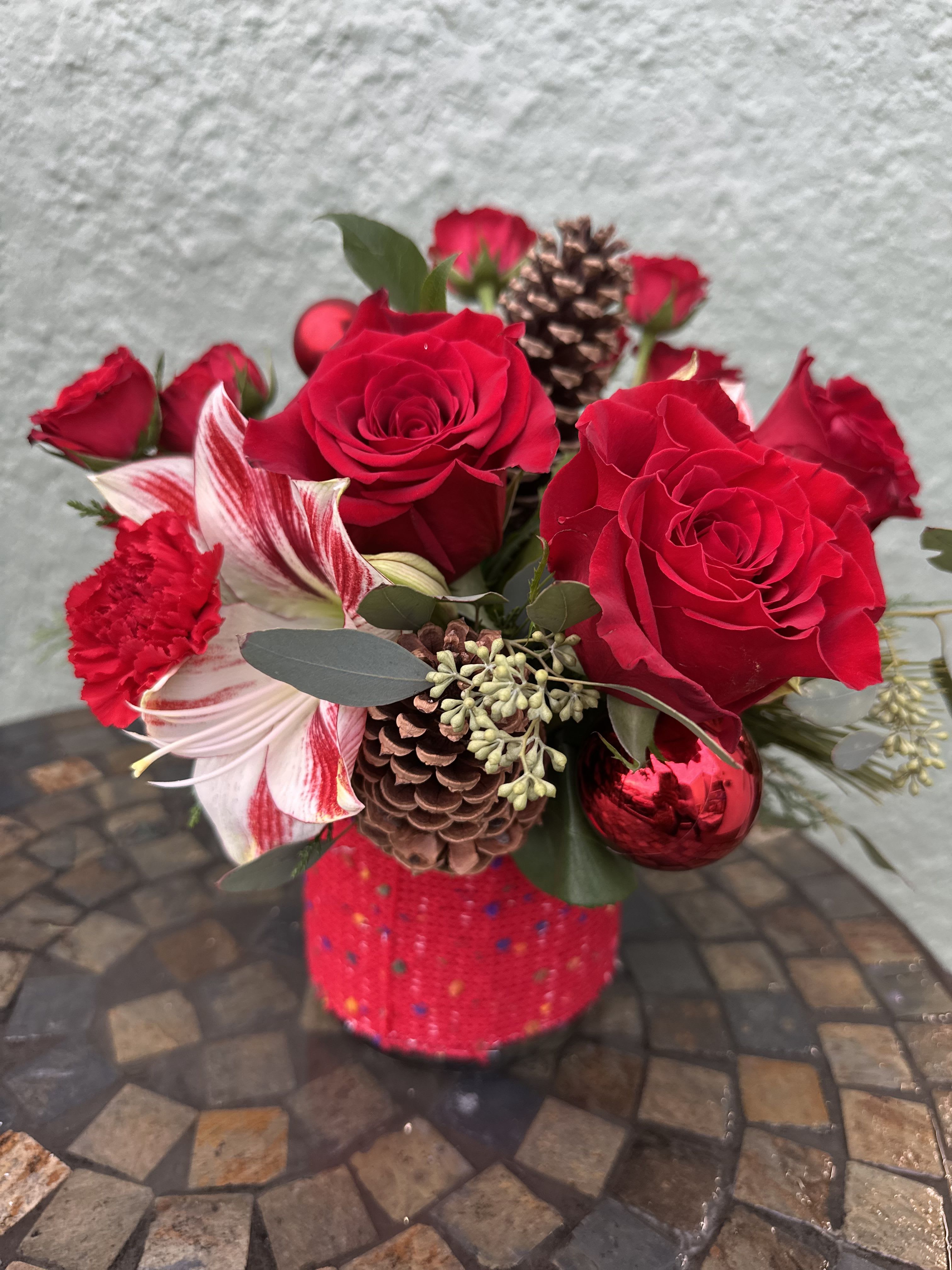 Yultide Cheer - Bring the warmth and joy of the season indoors with this classic and vibrant arrangement! Deep red roses and carnations are nestled among variegated Ambience amaryllis and lush seasonal greens, accented with natural pinecones and shimmering red baubles. The glass vase is wrapped with warm red sweater-like fabric. It's the perfect statement piece for your holiday table or a stunning gift to spread cheer!