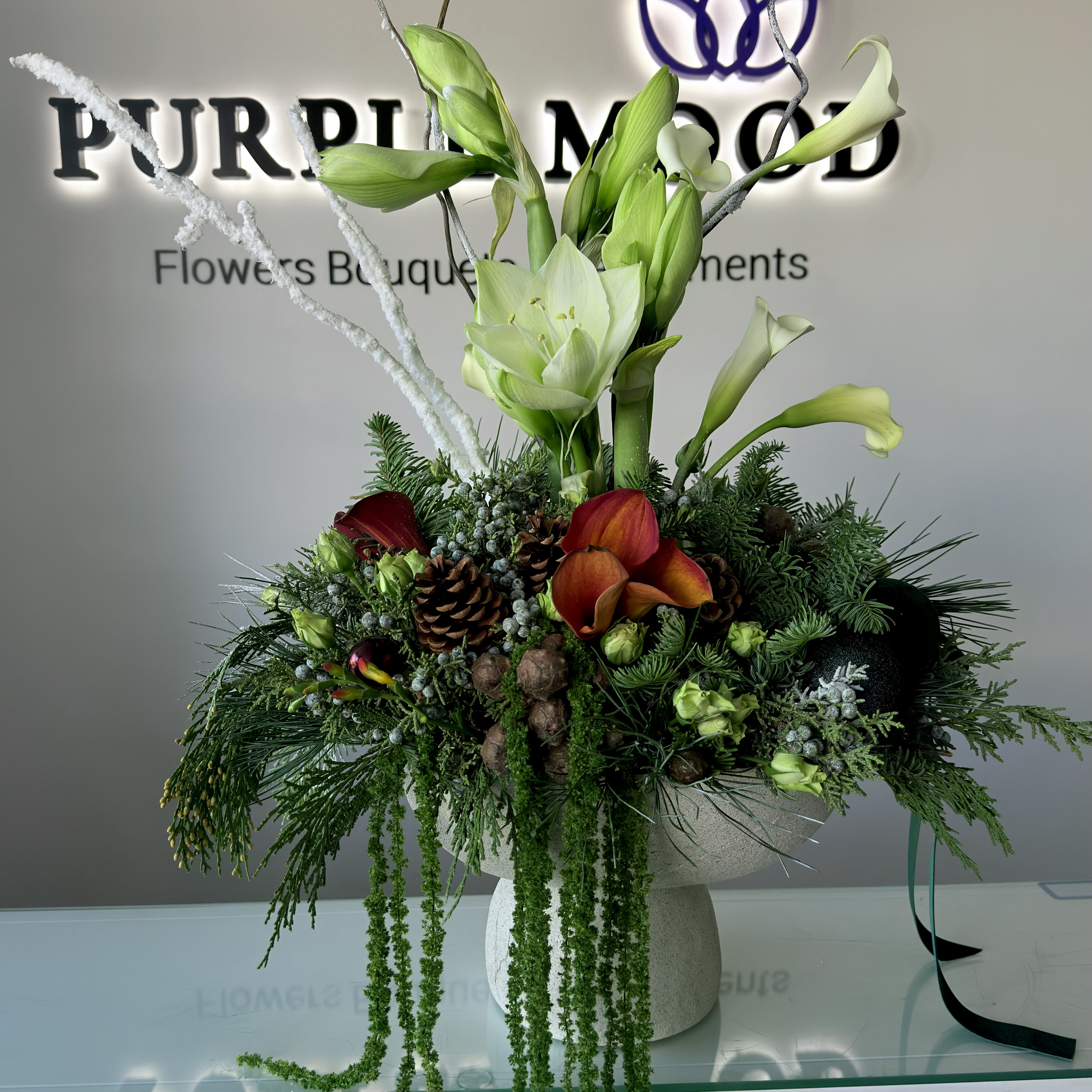 Evergreen Elegance Vase - The Evergreen Elegance Vase is a modern winter arrangement designed to bring the beauty of a serene holiday forest indoors. Featuring lush seasonal greenery, premium amaryllis stems, deep burgundy and amber calla lilies, natural pinecones, winter berries, and textural accents, this design combines rich color with organic sophistication.  Arranged in a contemporary stone-textured vase, this centerpiece offers a perfect balance of height, movement, and festive charm. Ideal for holiday tables, luxury gifting, or elevating any winter interior.  Perfect for: holiday celebrations, home décor, seasonal gifting, corporate and hostess gifts.  Flowers &amp; Elements Include: – Fresh amaryllis – Premium calla lilies – Seasonal evergreens – Pinecones &amp; winter berries – Textural accents  A The Evergreen Elegance Vase is a modern winter arrangement designed to bring the beauty of a serene holiday forest indoors. Featuring lush seasonal greenery, premium amaryllis stems, deep burgundy and amber calla lilies, natural pinecones, winter berries, and textural accents, this design combines rich color with organic sophistication.  Arranged in a contemporary stone-textured vase, this centerpiece offers a perfect balance of height, movement, and festive charm. Ideal for holiday tables, luxury gifting, or elevating any winter interior.  Perfect for: holiday celebrations, home décor, seasonal gifting, corporate and hostess gifts.