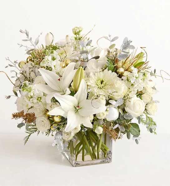 Fresh Frost Arrangement   - NEW As white and glimmering as freshly fallen snow, this arrangement captures the magic of the season in a most sophisticated way. White roses, Asiatic lilies, and other blooms are complemented with winter greenery accented with hints of gold and silver, along with gold and silver ball ornaments. It’s a wonderful way to add even more sparkle to the holidays.