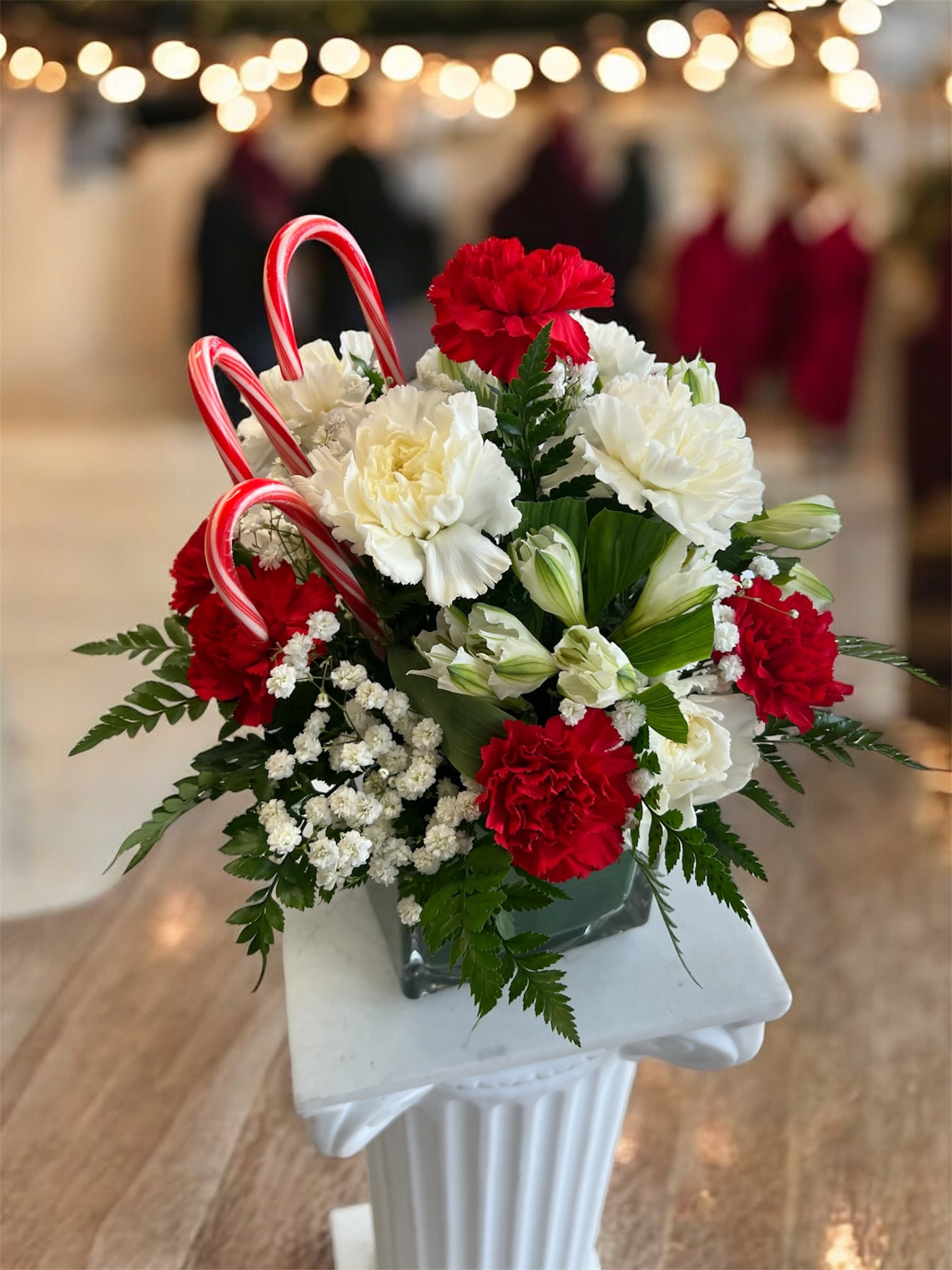 Candy Cane Delight  - Bring festive charm into any space with this delightful Christmas-inspired arrangement! Featuring vibrant red and creamy white carnations, fresh greenery, and delicate baby’s breath, this design captures the warmth of the season. Accented with playful candy canes for a sweet holiday touch, it’s perfect for gifting, decorating your home, or adding cheer to any holiday gathering. Arranged in a modern glass cube vase for a clean, elegant finish.  Lleva el espíritu festivo a cualquier lugar con este hermoso arreglo inspirado en la Navidad! Con claveles rojos vibrantes y blancos cremosos, follaje fresco y delicada nube, este diseño captura la esencia cálida de la temporada. Decorado con bastones de dulce que añaden un toque dulce y divertido, es perfecto para regalar, decorar tu hogar o alegrar cualquier reunión navideña. Presentado en un elegante florero cúbico de cristal.
