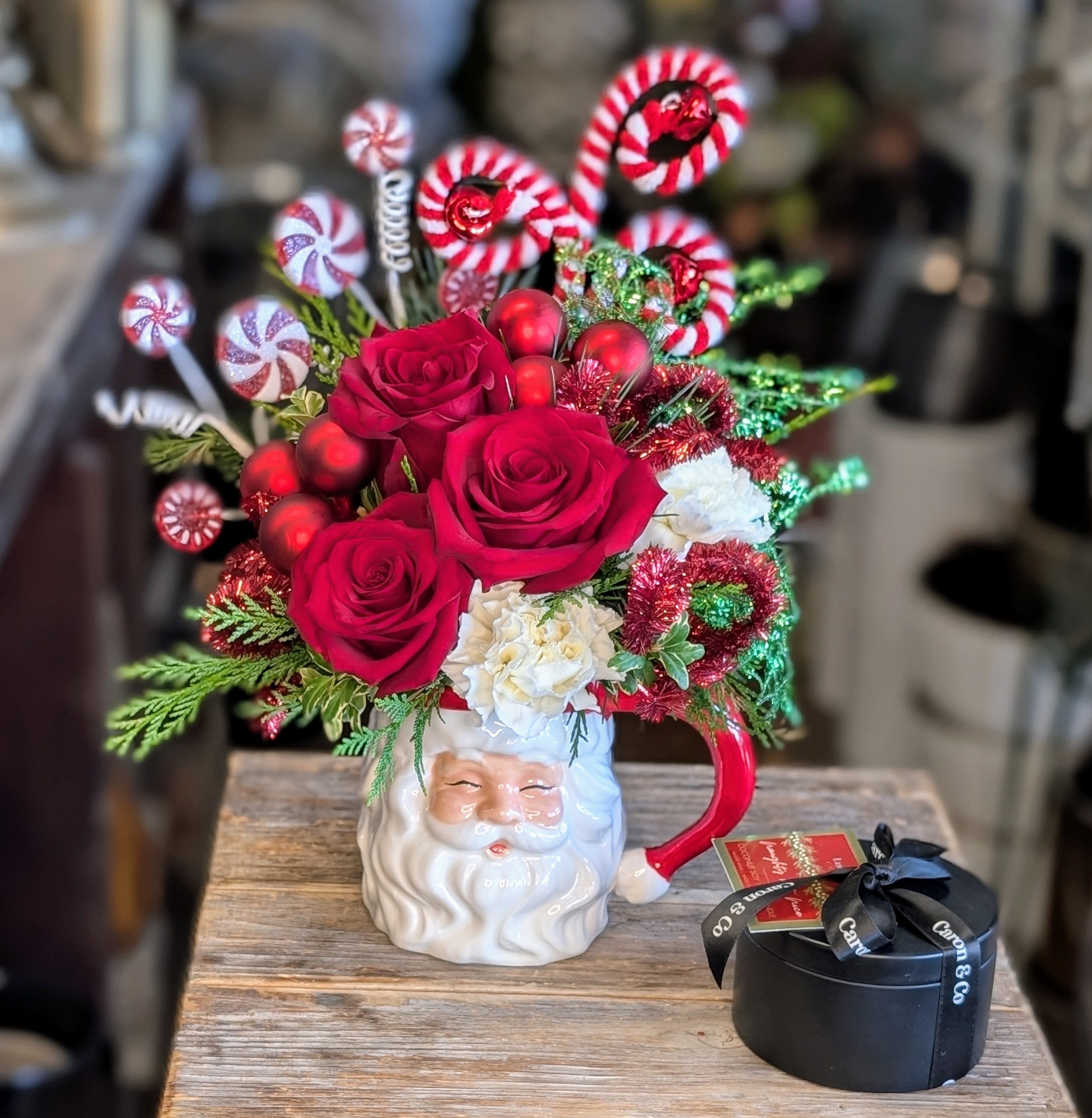Naughty But Nice With A Naughty But Nice Soy Candle - Naughty But Nice – A Playful Peppermint Holiday Arrangement Vibrant, festive, and full of cheeky charm, Naughty But Nice is a holiday showstopper designed by Windmill Floral Expressions in Las Vegas. Bursting from a vintage-inspired Santa mug, this lively arrangement features bold red roses, fluffy white blooms, and a joyful explosion of peppermint swirls, candy canes, and shimmering holiday baubles.  Glittered greenery, sparkly picks, and bright red ornaments give this design its over-the-top holiday magic—perfect for anyone who loves Christmas with a side of sass. Whether placed on a desk, kitchen counter, or holiday party table, Naughty But Nice instantly brings smiles and festive spirit.  Paired with a luxe candle, this arrangement is a fun, memorable way to celebrate the season—sweet, spirited, and just a little mischievous.  It is accompanied by a Naughty But Nice candle, a captivating blend of warm cinnamon and tonka and vanilla spices that capture the holiday season. It is a soy coconut wax with a wood wick, 5.5 oz candle with 30 + hours burn time.