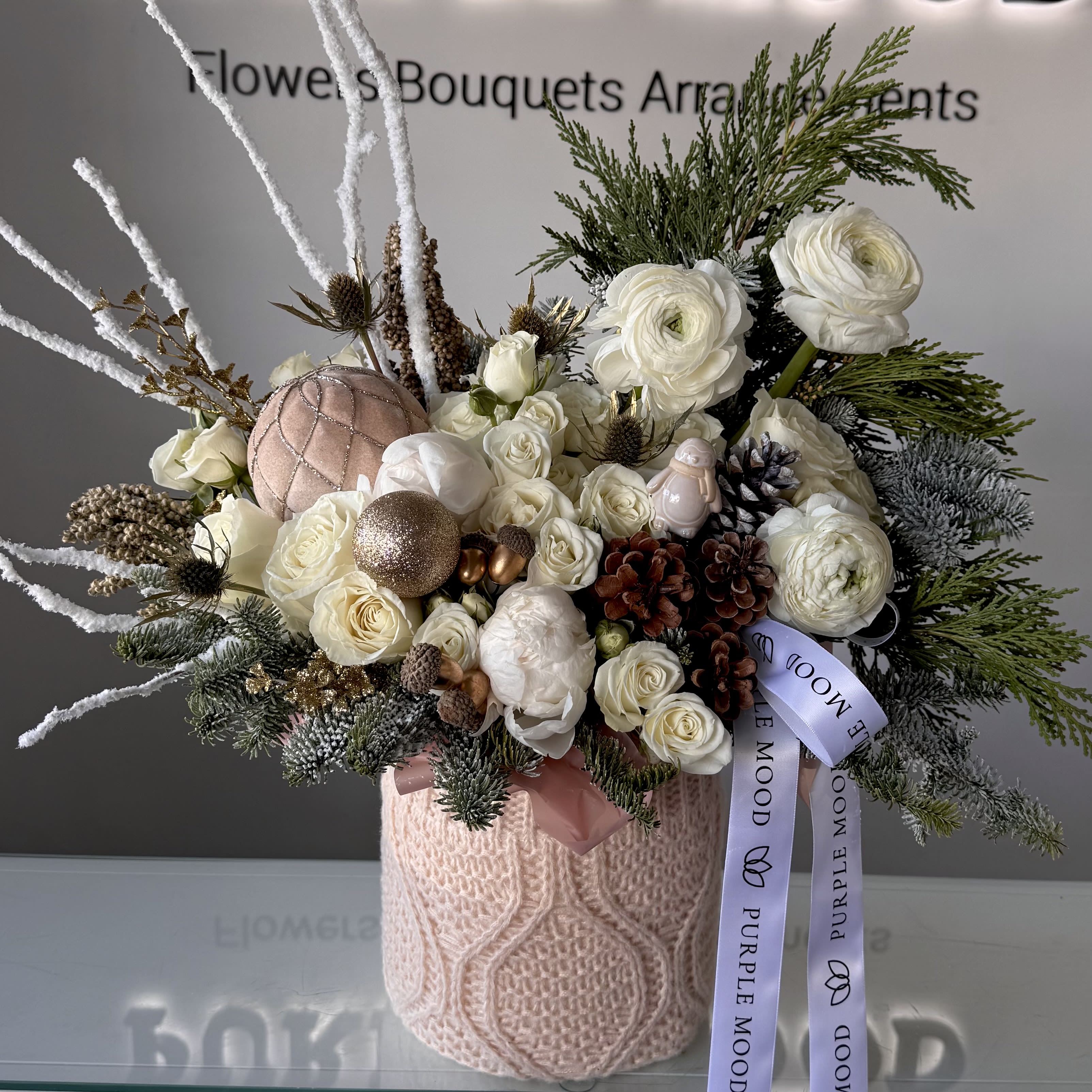 Snowy Knit Box - The Snowy Knit Box is a soft and elegant winter arrangement designed to bring warmth and festive charm into any space. Created in a cozy knit-textured vase, this piece blends creamy white blooms with delicate seasonal greenery, natural pinecones, and shimmering holiday accents.  Featuring premium roses, ranunculus, winter fillers, and carefully selected decorative ornaments, this arrangement captures the feeling of a peaceful snowy morning — gentle, bright, and full of holiday spirit.  Perfect as a holiday gift, home décor accent, or a stunning centerpiece for winter gatherings.  Perfect for: seasonal gifting, holiday décor, home styling, corporate gifting.
