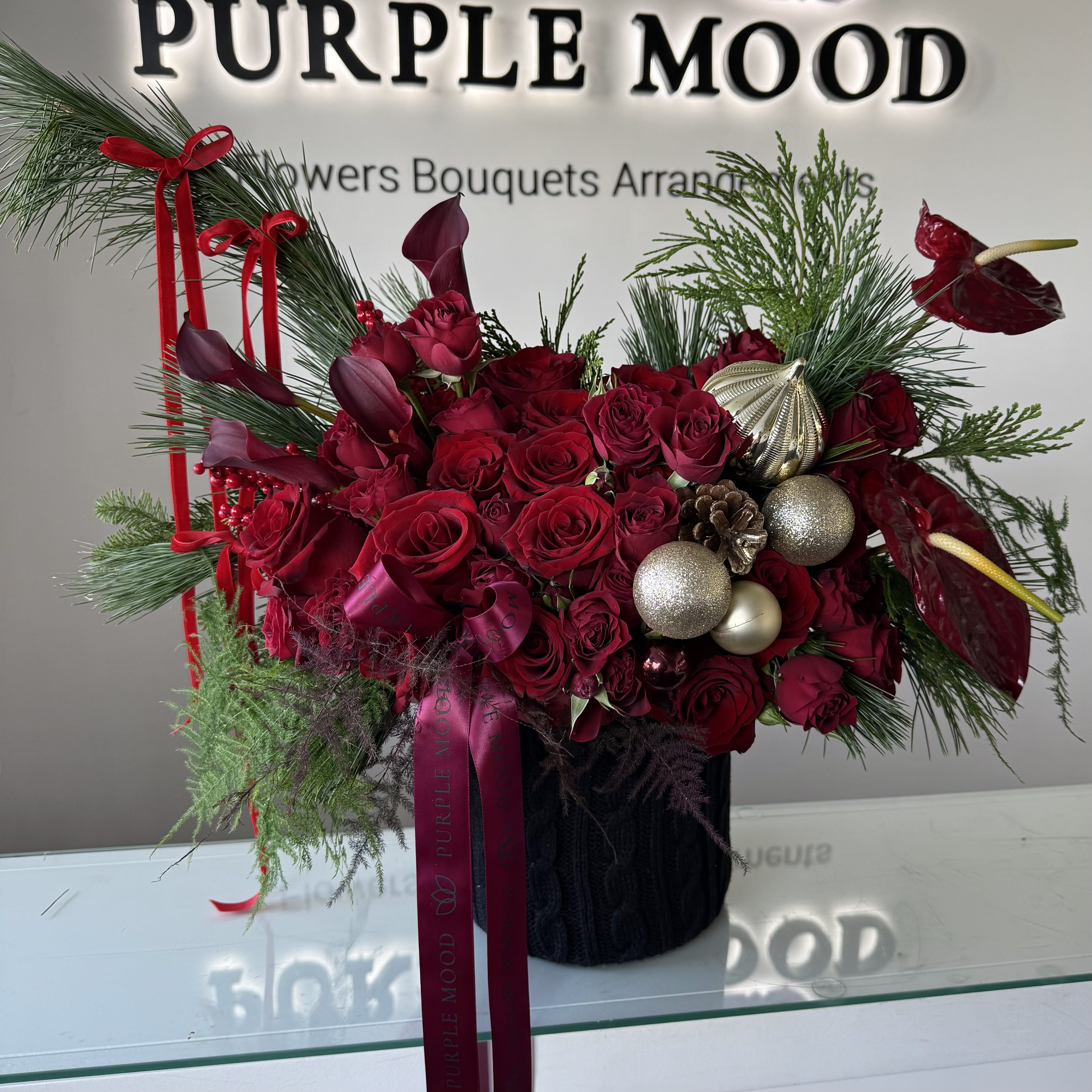 Red Knit Celebration Box - The Red Knit Celebration Box is a luxurious holiday arrangement bursting with rich crimson tones and festive winter textures. Designed in a premium knit-textured vase, this piece showcases an abundance of velvety red roses, deep burgundy calla lilies, elegant anthuriums, and seasonal greenery for a dramatic yet refined holiday statement.  Golden ornaments, pinecones, and winter berries add a touch of sparkle, making this arrangement the perfect centerpiece for holiday gatherings, upscale gifting, or elevating any seasonal décor.  Bold, celebratory, and full of festive charm — this design brings the magic of the holidays to any room.  Perfect for: holiday celebrations, luxury gifting, home décor, corporate events.
