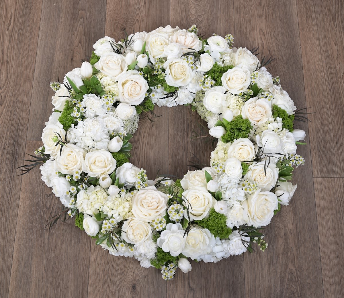 Wreath-White  -  Graceful, serene, and deeply elegant, Wreath-White  is a timeless expression of love and remembrance. Lush white blooms are artfully arranged in a full, layered circle, creating a soft and peaceful silhouette that symbolizes purity, honor, and everlasting connection. Delicate textural accents and natural greenery add gentle movement and depth, giving the design a modern, refined presence suitable for memorial services, celebrations of life, or heartfelt condolences.  Please Note: Our arrangements are handcrafted, and no two pieces are ever identical. Variations in flower types, color tones, and exact placement will occur based on daily availability. We do not create factory-duplicate designs — instead, we recreate the style, palette, and overall feel of the arrangement while ensuring every piece is fresh, beautiful, and premium.