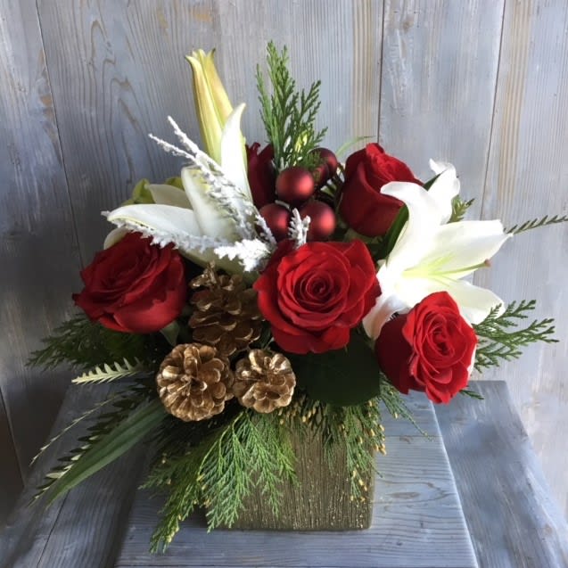Christmas Spirit - Arrangement in a white  ceramic container filled with; red roses, pine greenery, lilies, red ornaments, and pine cones. ( Deluxe includes cymbidium orchids.)