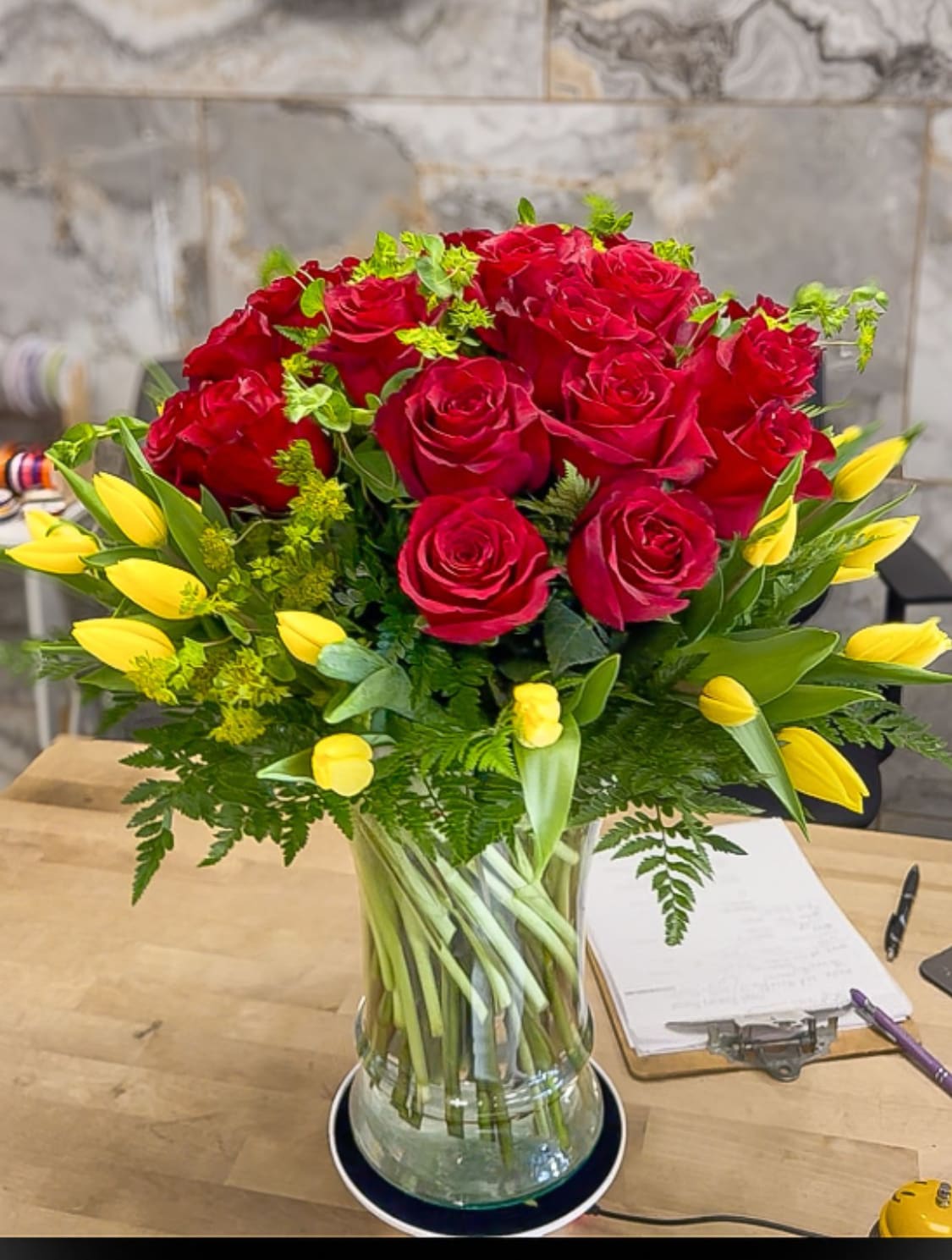 Rosalia - A lush, dramatic bouquet in a clear glass vase, centered with two dozen deep crimson red roses and punctuated by twenty sunny yellow tulips. The vibrant red and yellow bloom mix is framed by glossy greenery and delicate ferns, adding depth and a touch of elegance. The clear glass vase keeps the look timeless and fresh, making this arrangement a refined centerpiece ideal for gifting, events, or brightening any space with warm, celebratory energy. Deluxe 36 roses 30 tulips 