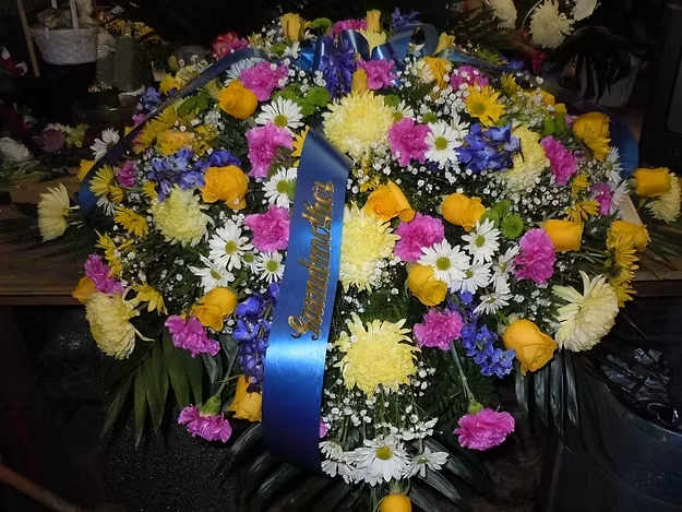 C2 Casket Spray - Casket spray with a mix of yellow fuji mums, white and yellow daisies, ferida carnations, blue delphinium, yellow roses, baby's breath, and greens.   Banners - $15.00 + extra  Substitutions may apply, pending flower availability.