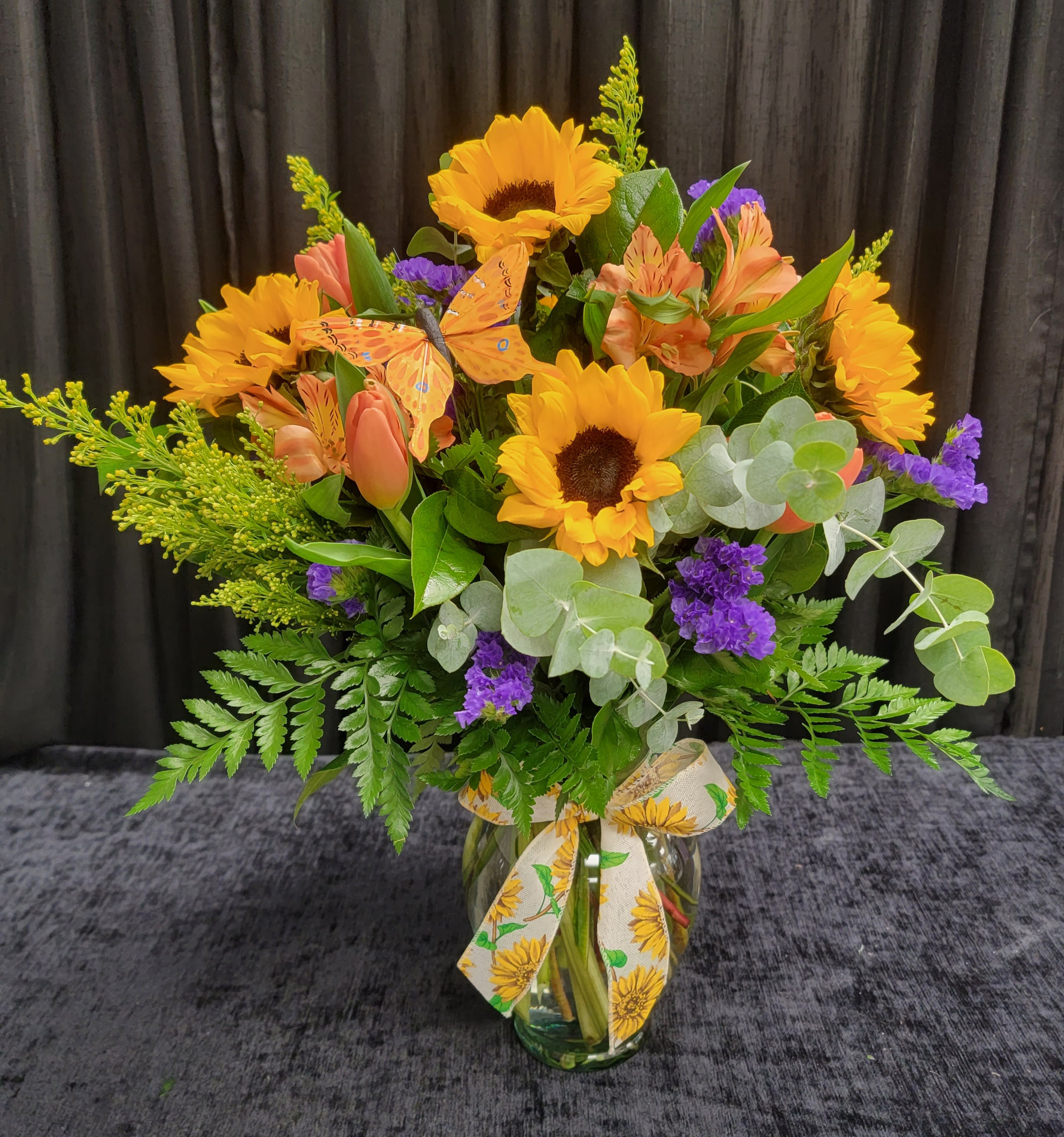 Butterfly Magic - A bright beautiful vase filled with sunflowers, tulips, filler flowers and a butterfly. Colors may vary slightly.