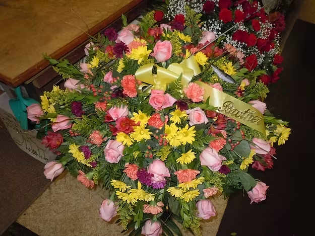 C8 Casket Spray - A mixture of light pink rose, orange carnations, yellow and purple daisies, solidago, red hypericum, myrtle, burgandy alstro, and greens.   Banner - $15.00 + extra  Substitutions may apply, pending flower availability.