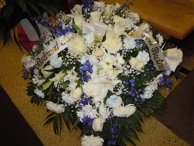 C11 Casket Spray - A mixture of white fuji mums, white roses, white lilies, white carnations, white daisies, dark blue delphinium, greens, and baby's breath.  Includes  bow.  Banner - $15.00 + extra  Substitutions may apply, pending flower availability.