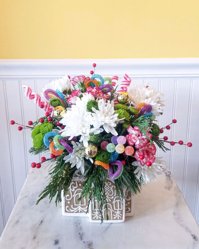 Gingerbread Dreams - This delightful arrangement captures the sweet charm of the holiday season with its gingerbread-themed accents, festive red and white blooms, and lush greenery. Perfect for adding a touch of magic and warmth to your holiday decor or for gifting to loved ones. approx 14 X 13