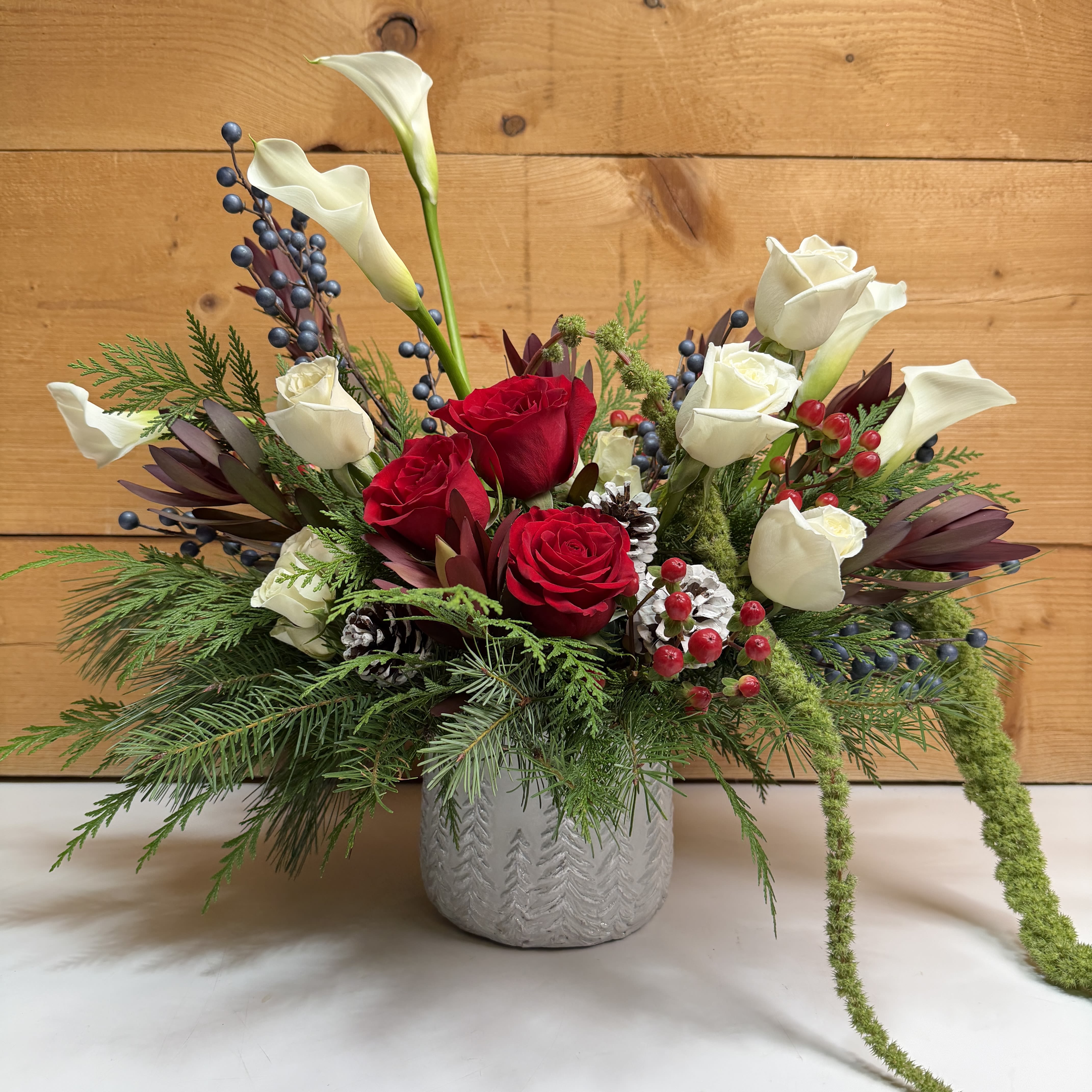 Into The Woods - Into The Woods is a magnificent display of elegant blooms designed to give your holiday decor a sense of sophistication and class. This design includes calla lilies, roses, safari sunset, and more, joined with delicate, faux blue berries, in a gorgeous white pine tree themed container. This design is a stellar way to decorate for the holidays in a glamorous way, combining style and winter into one phenomenal piece.  Dimensions approx. L 20” x H 21”