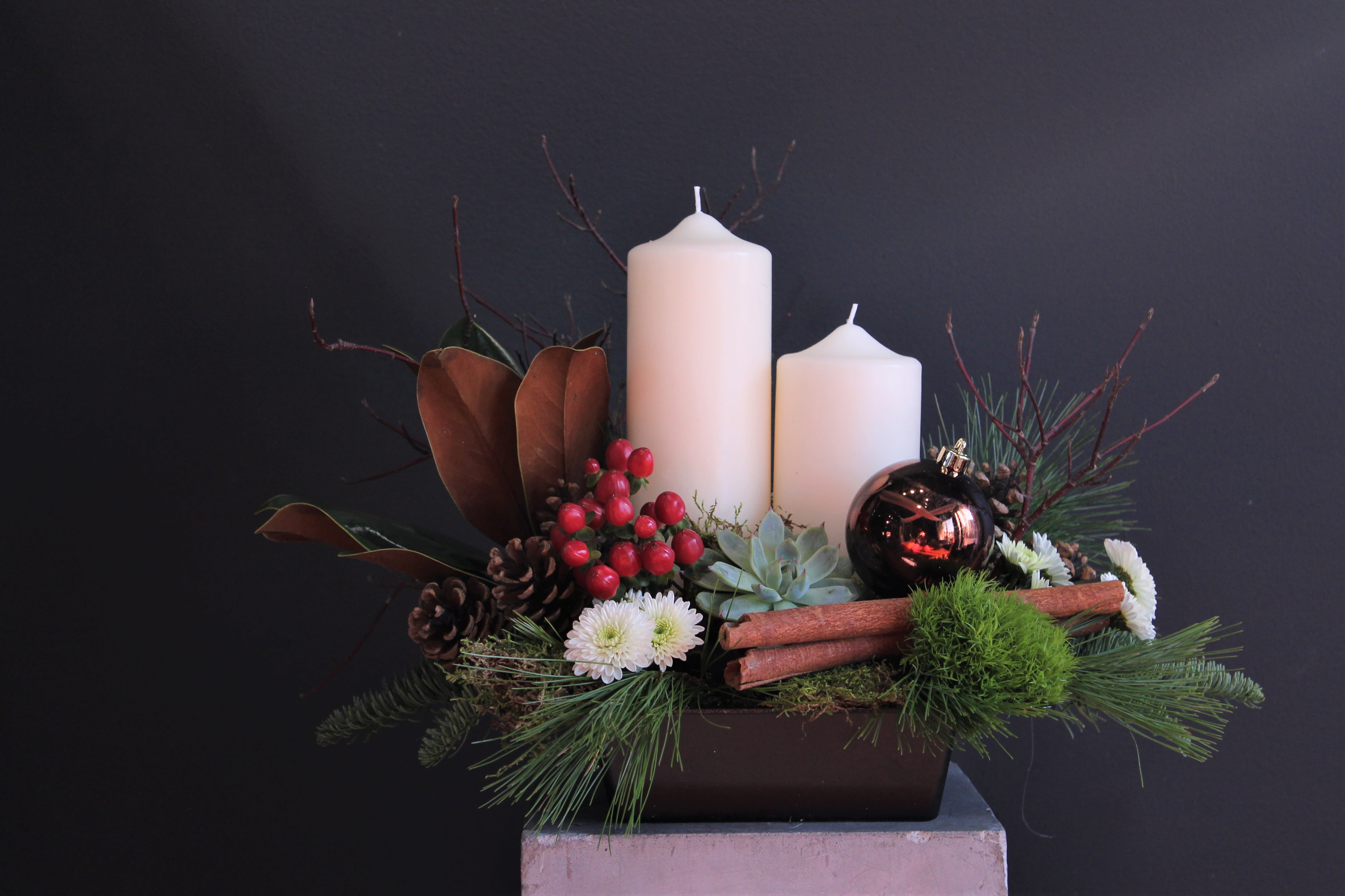 Cinnamon Spice Everything Nice - This arrangements warm, festive, and elegantly rustic design captures the cozy charm of the holiday season in a modern centerpiece.  Nestled in a sleek dark rectangular container, this arrangement features two ivory pillar candles of differing heights, perfect for creating a soft, inviting glow. Around the candles, a lush mix of evergreen sprigs, magnolia leaves, and pinecones evokes the scent and texture of a winter forest.  Touches of bright red berries add a pop of cheerful color, complemented by fresh white chrysanthemums and a succulent rosette that lends a contemporary twist. Two aromatic cinnamon sticks rest among the greenery, bringing warmth and seasonal spice to the design. A shiny bronze ornament adds a touch of holiday sparkle and refinement.  The overall effect is earthy yet elegant, blending natural elements with subtle festive accents — the perfect centerpiece for a Christmas table or a welcoming holiday display.