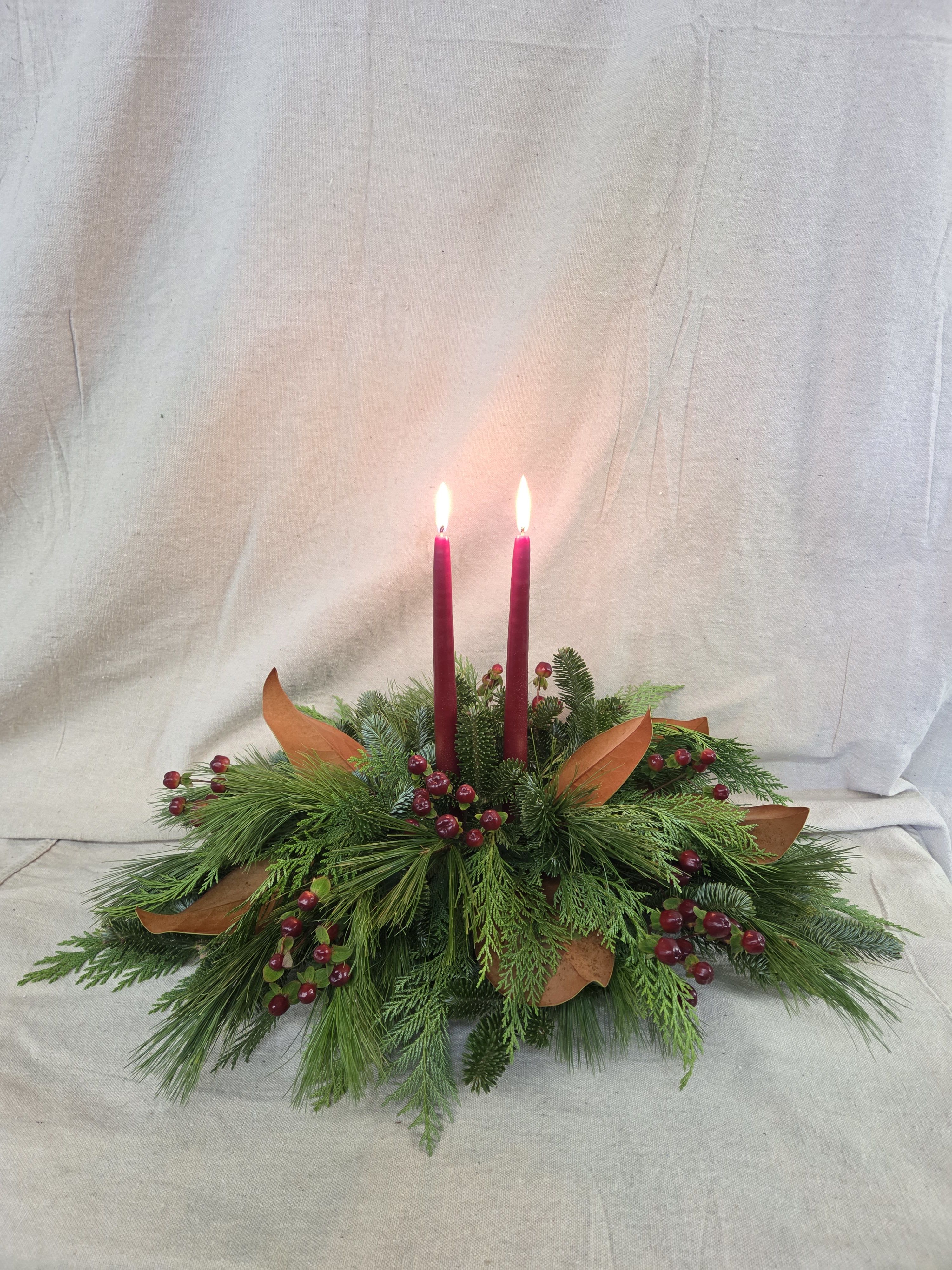 Winter Gathering Centerpiece - Bring timeless holiday charm to your table with this lush Christmas green centerpiece. Layered with fresh evergreens—like cedar, fir, and pine—this arrangement is accented with subtle seasonal touches and finished with tall taper candles that cast a warm, inviting glow.