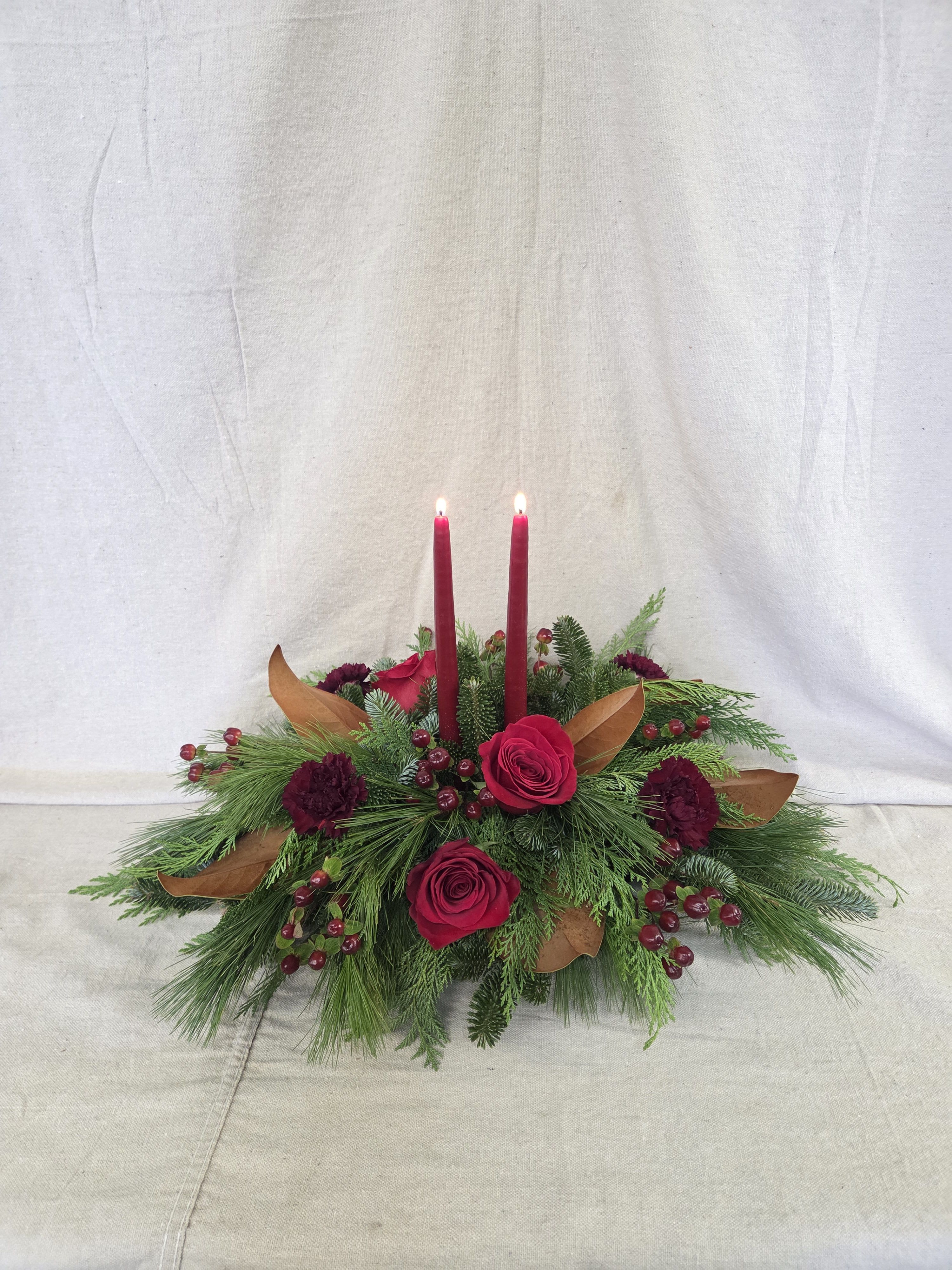 Holiday Charm Centerpiece - This timeless Christmas centerpiece blends lush Christmas greens with velvety red roses for a look that’s rich, festive, and refined. Tall taper candles rise above the arrangement, casting a soft, romantic glow that enhances every holiday gathering.