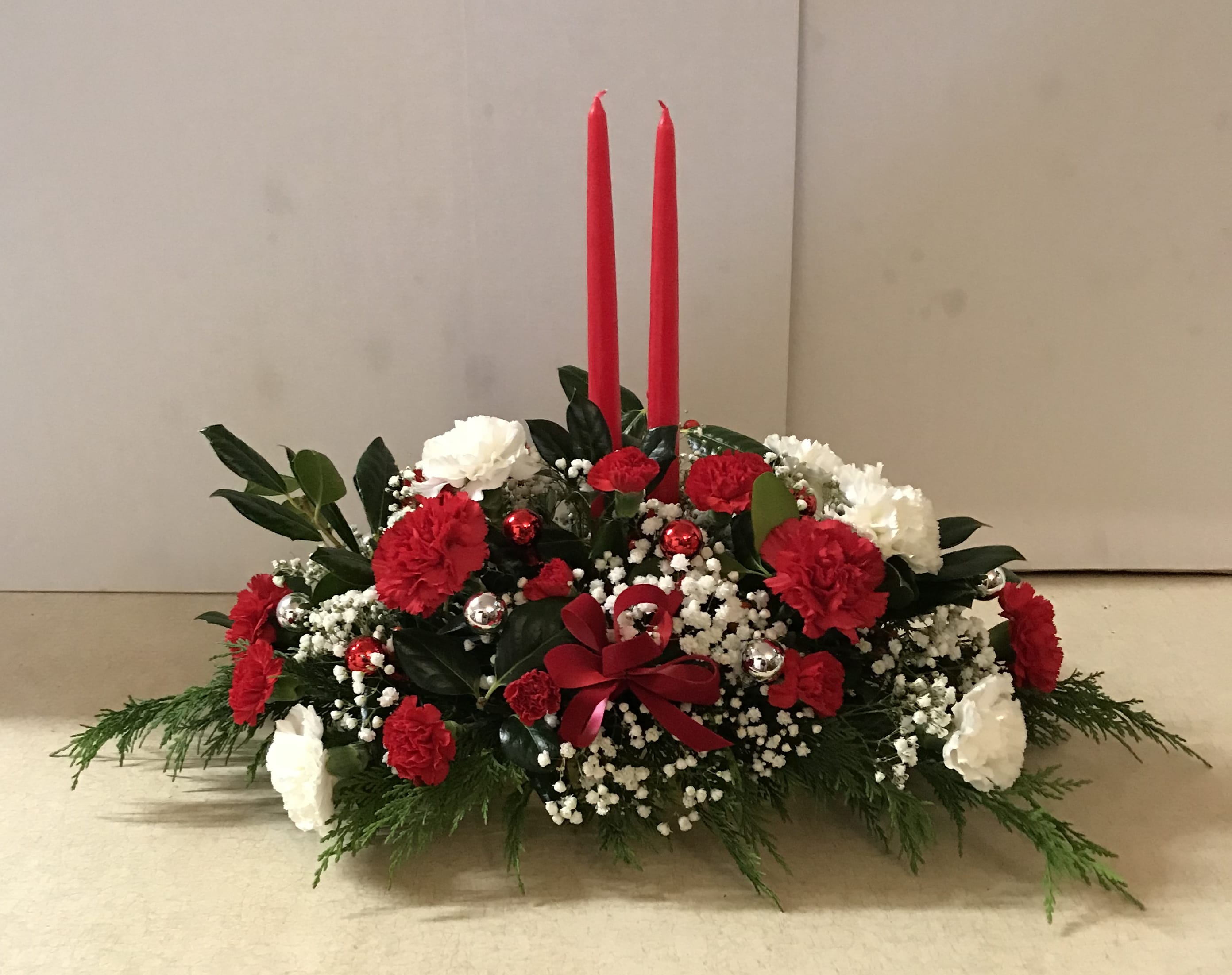 Christmas Candles and Holiday Flowers - This oblong centerpiece sets inside a gold compote and is filled with holiday greenery, red carnations, red-mini carnations, white carnations, baby's breath, and fresh holly.  There are two red 15" candles included, as well as red and silver Christmas balls and a red bow.  The pictured design sells for $75.00 and is the deluxe choice.  It makes a lovely centerpiece for any holiday setting.  Get yours now so you can enjoy it throughout the festive season!