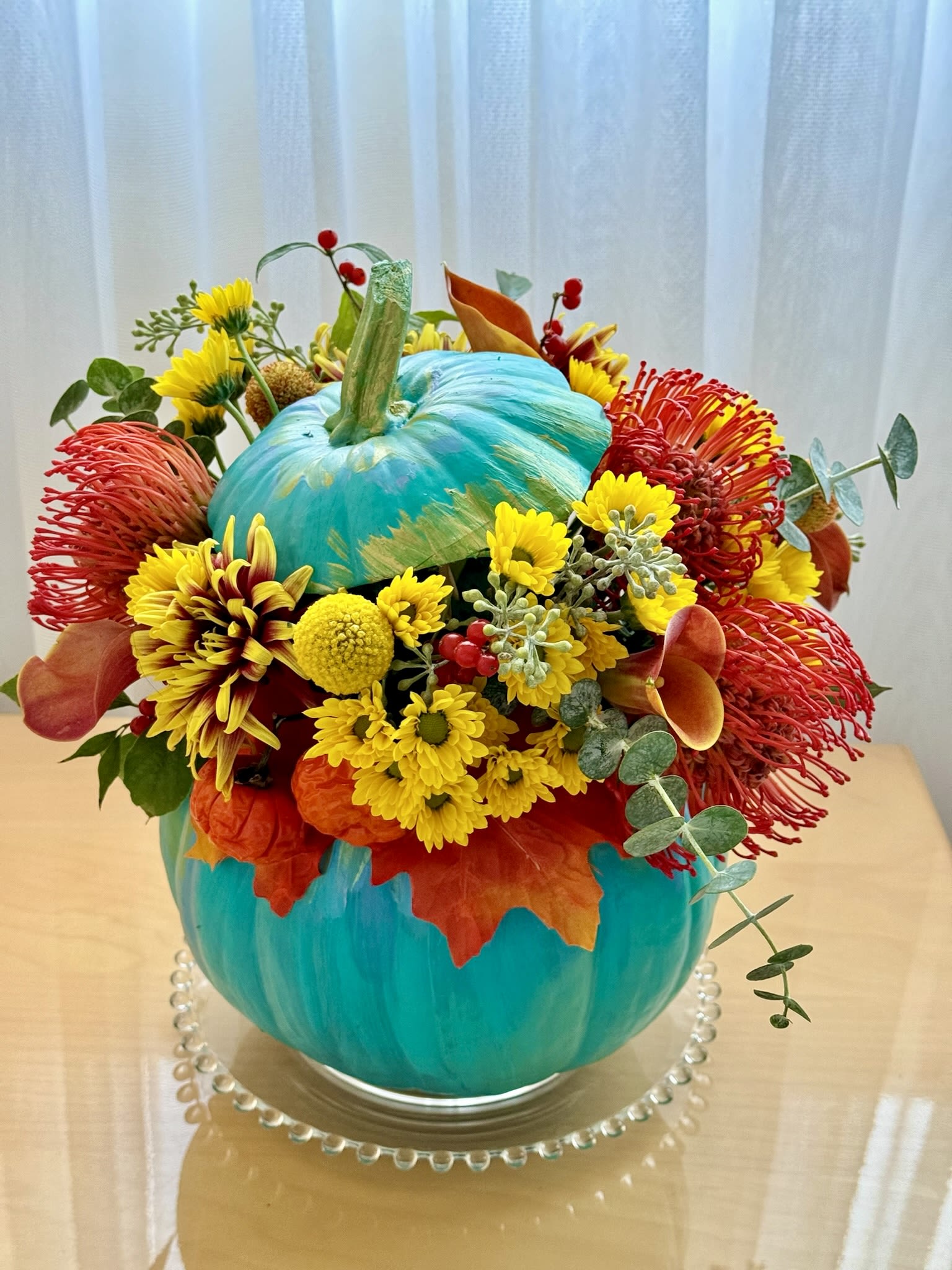 Pumpkin Fiesta - This vibrant and playful fall arrangement is designed in a stunning turquoise pumpkin vase, offering a bold twist on classic autumn décor. The arrangement features a striking mix of yellow chrysanthemums, red Leucospermum (pincushion flowers), orange calla lilies, and bright billy balls, accented with fresh eucalyptus and red berries. The rich hues of red, yellow, and orange evoke the warmth of the season, while the turquoise pumpkin adds a modern and whimsical touch. Perfect for brightening up any space, this arrangement is a cheerful and colorful addition to your fall celebrations or as a unique centerpiece.   Mums Pin Cushion Lilly Calla Craspedia  Height 11 inch Width 9 inch