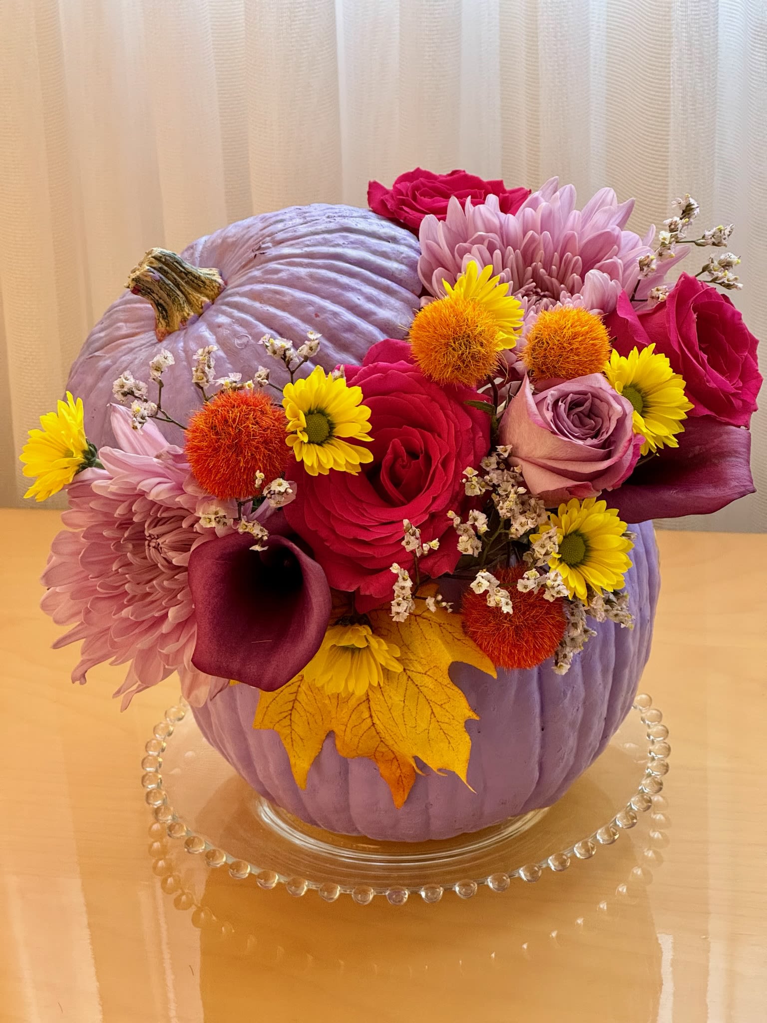 Lavender Pumpkin Delight - This eye-catching floral arrangement is designed in a bold lavender pumpkin vase, perfect for a unique and colorful autumn centerpiece. The arrangement features vibrant magenta roses, soft purple chrysanthemums, and dark purple calla lilies, contrasted beautifully with bright orange textured accents like billy balls and orange pom-poms. Delicate white filler flowers and golden autumn leaves add a seasonal touch, while the unexpected lavender pumpkin brings a modern twist to traditional fall décor. This arrangement is ideal for those looking to add a playful pop of color to their fall festivities or home décor.  Roses Calla Lilly Chrysanthemum  Height 11 inch Width 9 inch