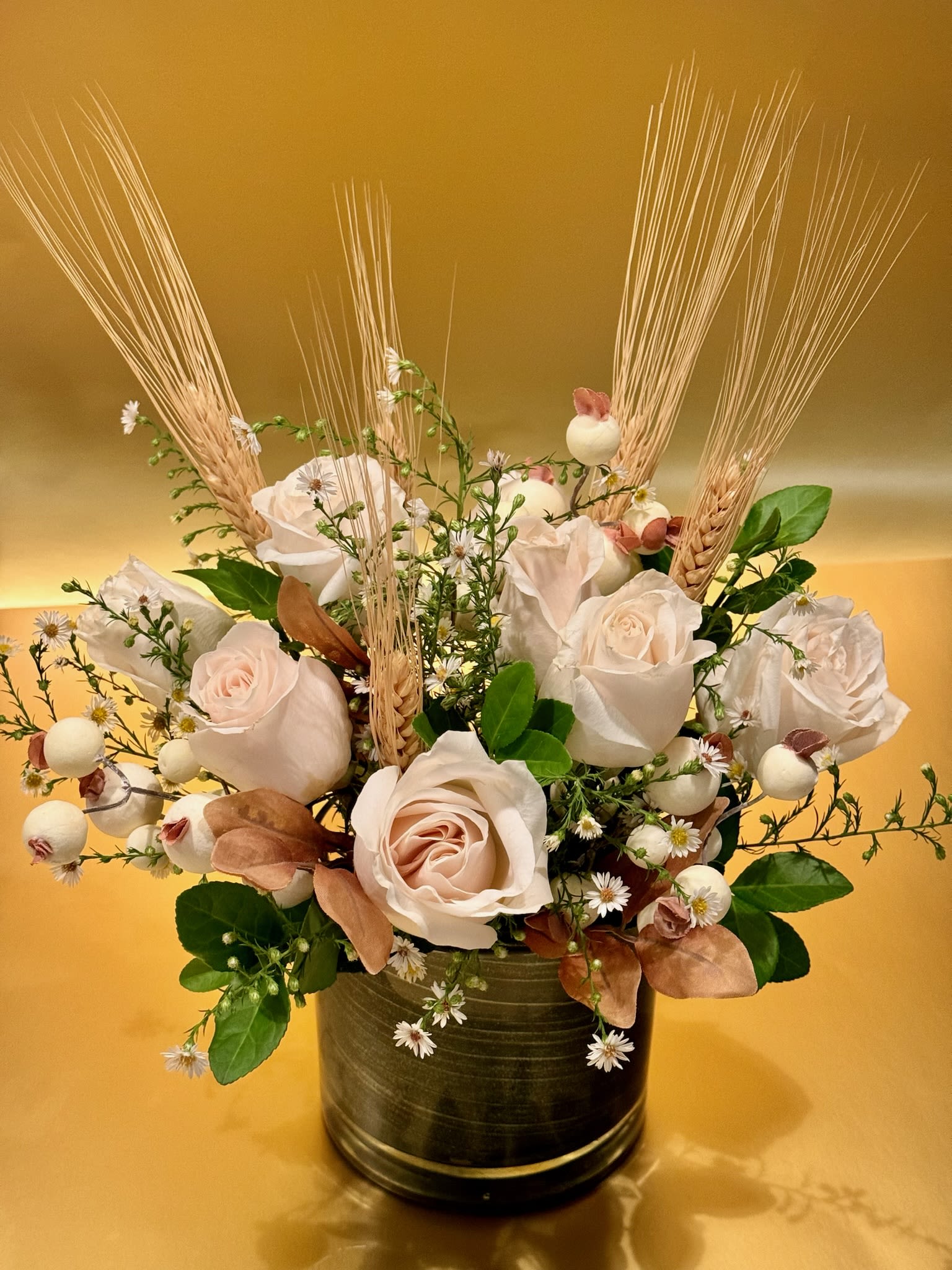 Autumn Rose Bouquet - This elegant and timeless arrangement features creamy white roses as the centerpiece, beautifully accented by soft greenery, delicate white waxflowers, and small white berries. Stalks of golden wheat add a rustic, autumnal touch, making this bouquet a perfect blend of natural beauty and seasonal charm. Designed in a sleek glass vase with a dark green wrap, this arrangement exudes sophistication, while subtle natural elements evoke the warmth of the harvest season.  Ideal for weddings, special occasions, or as a refined decorative piece, this arrangement brings a peaceful and graceful ambiance to any setting.   Roses Wheat Stalks Wax Flowers  Height 14 inch Width 9 inch