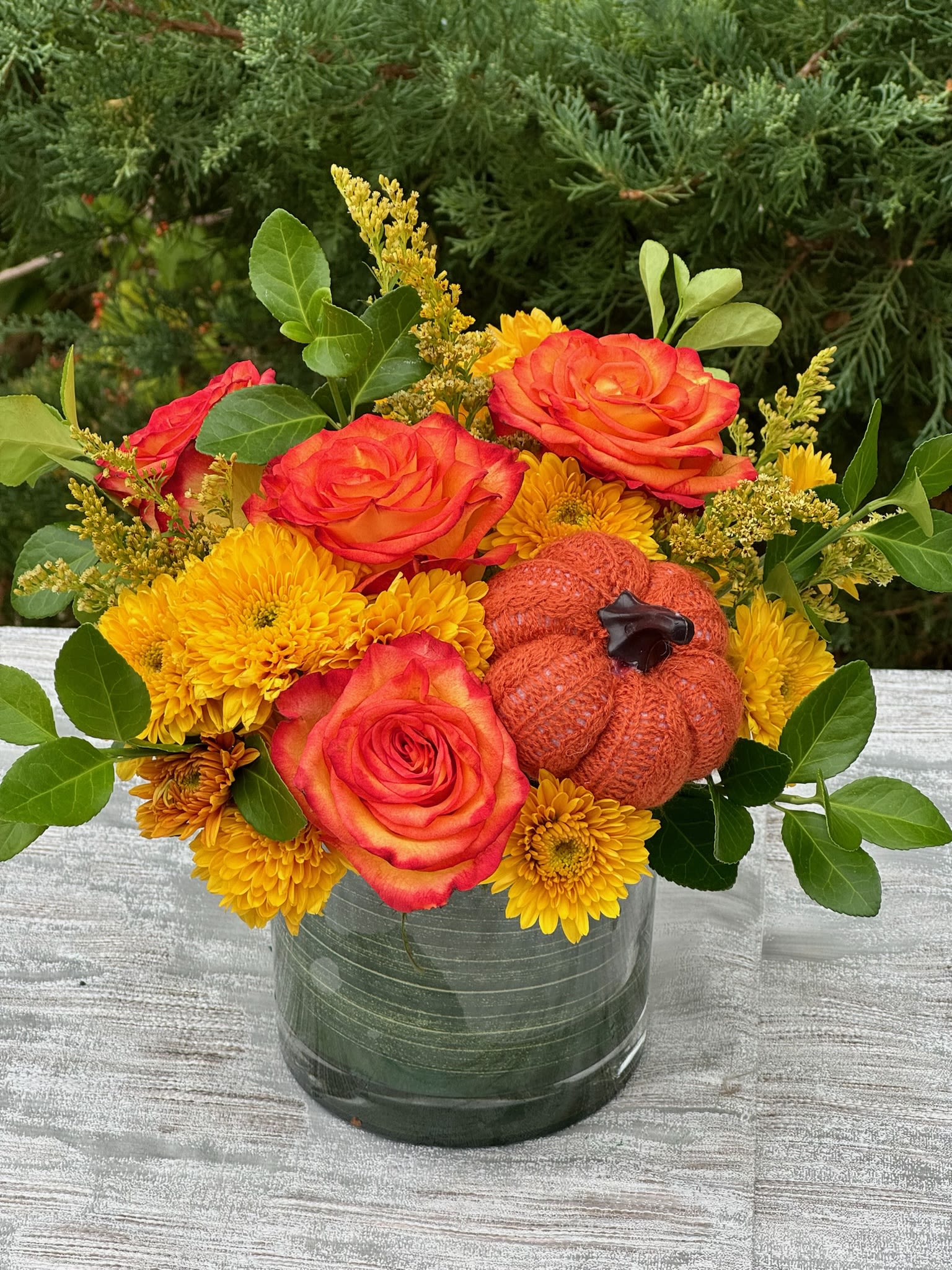 Pumpkin Spice Bouquet - The perfect floral arrangement to celebrate the warmth and colors of fall! This bouquet features bold orange roses and cheerful yellow mums, complemented by rich greenery and a charming knitted pumpkin accent. Designed in a sleek glass vase, it brings the cozy, rustic vibes of autumn straight to your home or event.  Whether you're gifting or decorating for the season, the Pumpkin Spice Bouquet adds a festive and welcoming touch.  Roses Mums Pumpkin  Height: 11 inch Width: 9 inch