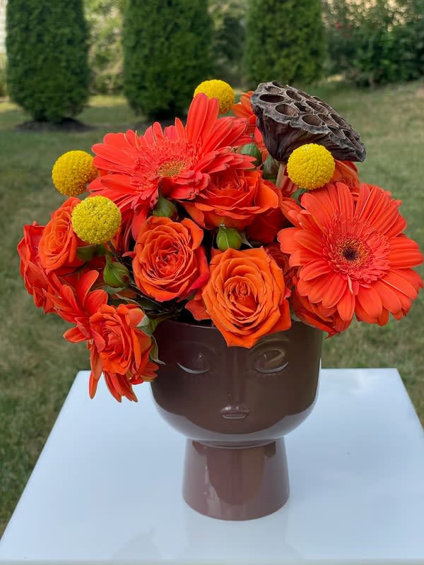 Autumn Expressions - This artistic bouquet features bright orange Gerbera daisies, vibrant roses, and yellow Craspedia, all beautifully arranged in a unique head-shaped vase with expressive facial features. The bouquet is accented with a dried lotus pod for an earthy, textured element, creating a playful yet elegant statement piece.  Perfect for art lovers or anyone looking to brighten up their space with a creative floral display.  Petit Roses Gerber Fall Accents  Height: 12 Inch Width: 9 Inch