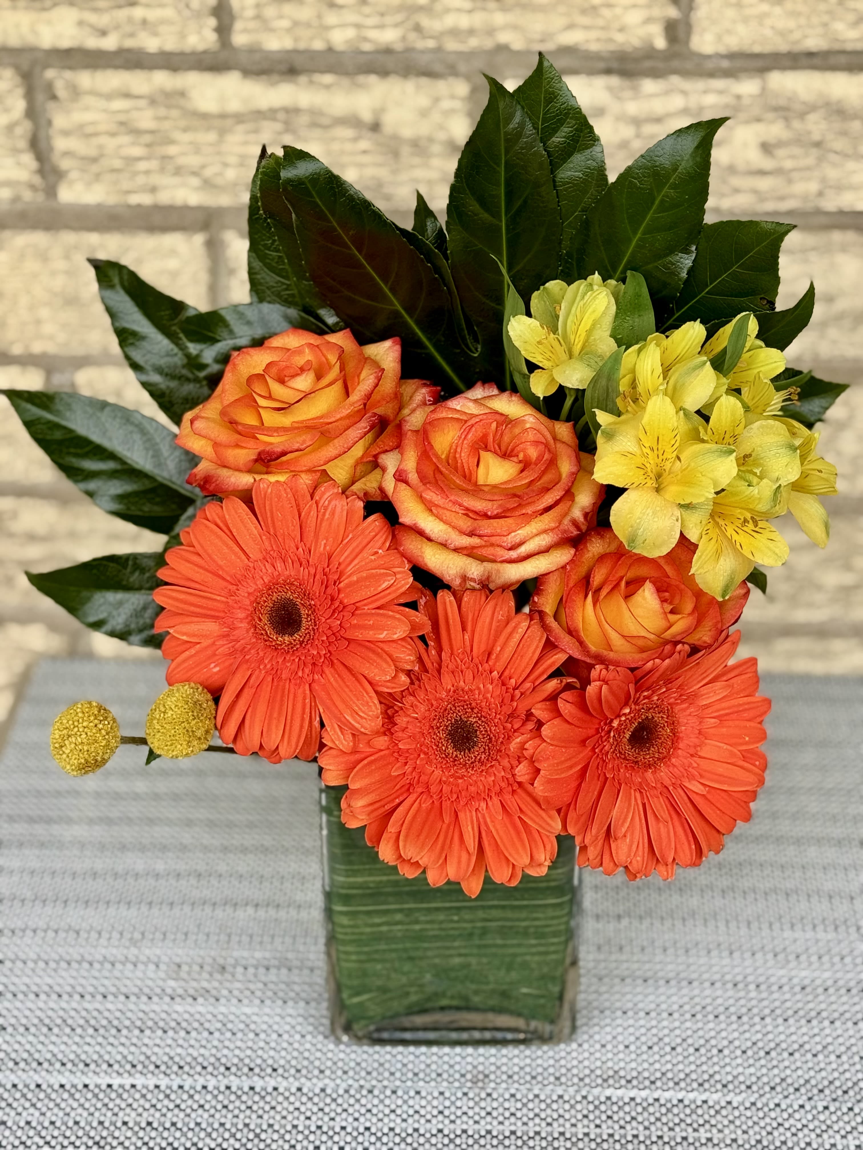 Orange Glow Bouquet - This bouquet is a celebration of the lively hues found in citrus fruits, exuding a sense of joy and radiance. The combination of these three blooms creates a harmonious and eye-catching display, making the &quot;Citrus Glow Bouquet&quot; a perfect choice for occasions that call for a burst of energy and a vibrant expression of happiness.  3 Orange Roses Orange Gerbera Craspedia  Height 16 in Width 15 in