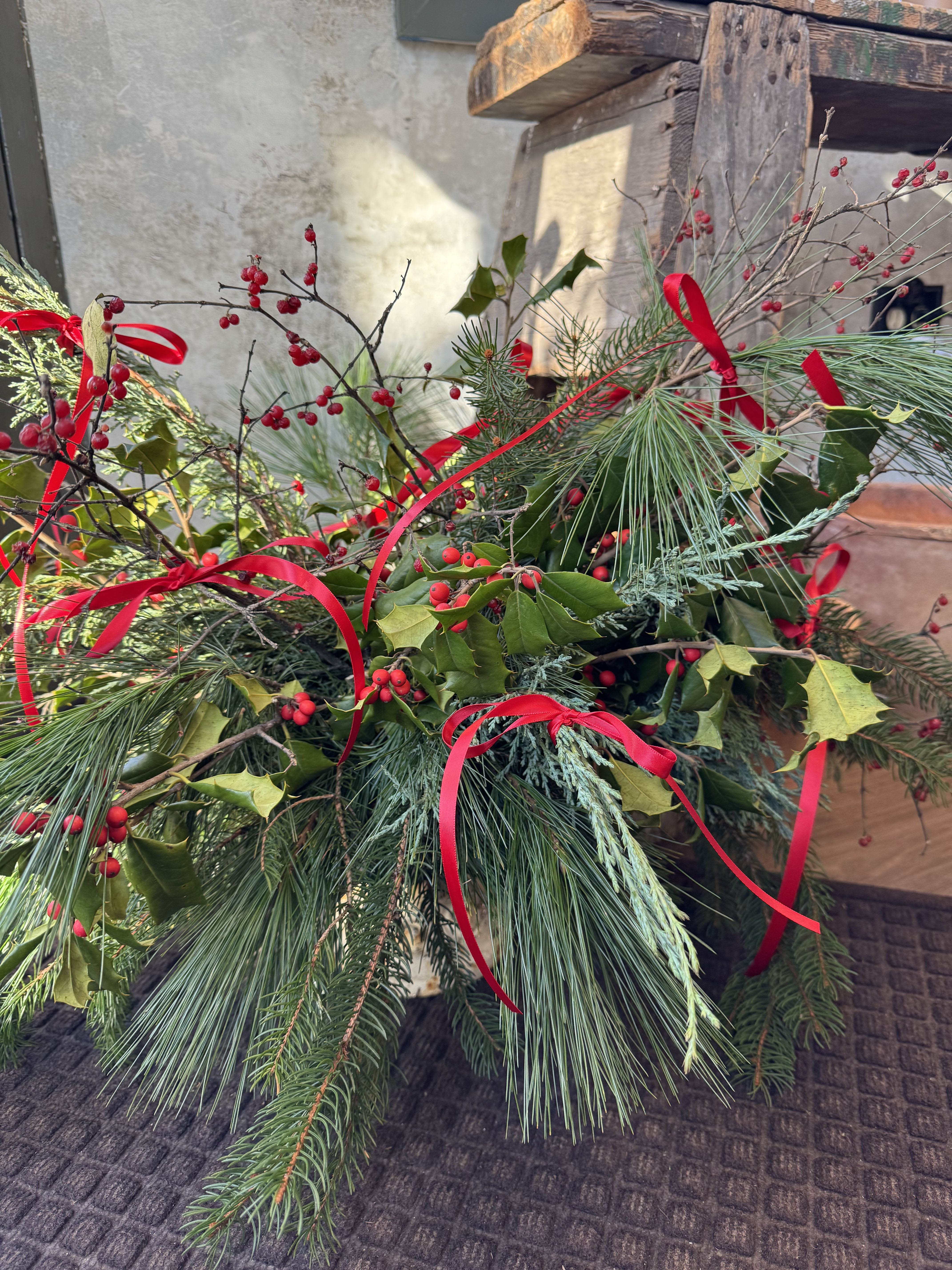 Holiday Wonderland Centerpiece - Our Holiday Wonder Centerpiece is a festive celebration of winter greens- full, textural, and arranged to feel like a little forest moment brought indoors. A mix of seasonal evergreens and winter branches is artfully styled into a long, low centerpiece that looks beautiful on dining tables, buffets, or entryways. Multiple ribbons in your choice of red or white are woven throughout the greens, adding a soft, cheerful touch without overwhelming the natural beauty of the foliage. Please specify ribbon color preference before checkout.