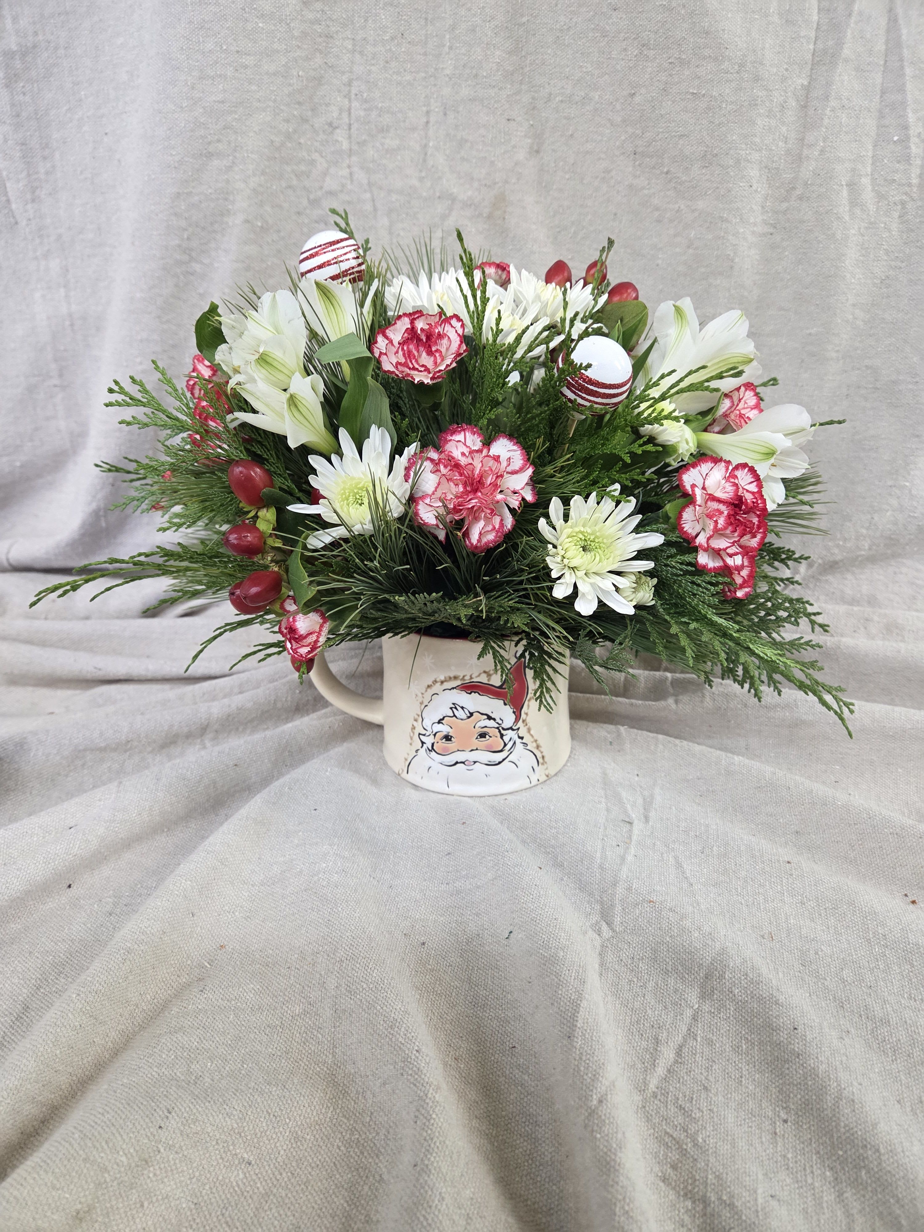 Winter Wishes in a Mug - This cheerful holiday arrangement brings a cozy touch of Christmas charm to any space. Designed in a festive seasonal mug, it features a delightful mix of fresh blooms, bright holiday accents, and lush winter greenery. Pops of red and white create a classic Christmas palette, while playful peppermint-inspired details add a sweet, whimsical touch. Perfect for gifting, decorating a desk or countertop, or spreading a little extra holiday joy throughout the season.