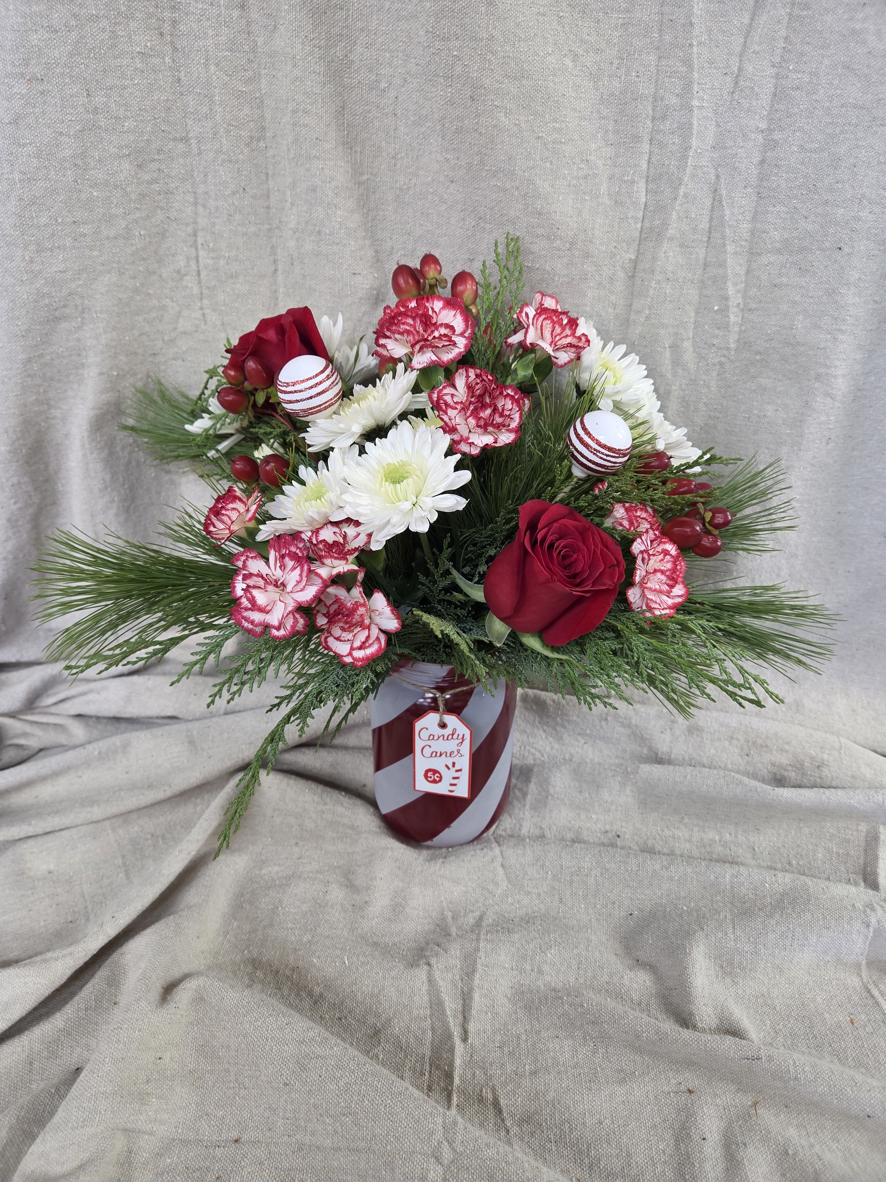 Peppermint Lane Bouquet - This festive holiday arrangement captures the sweetness and charm of the season. Designed in a striped candy-cane–inspired container, it features a cheerful mix of fresh seasonal blooms in classic red and white tones. Lush winter greenery adds fullness and texture, while playful peppermint accents bring a whimsical touch of Christmas magic. Perfect for brightening a holiday table, gifting to someone special, or adding a joyful pop of color to any room, this arrangement is a delightful celebration of the season.