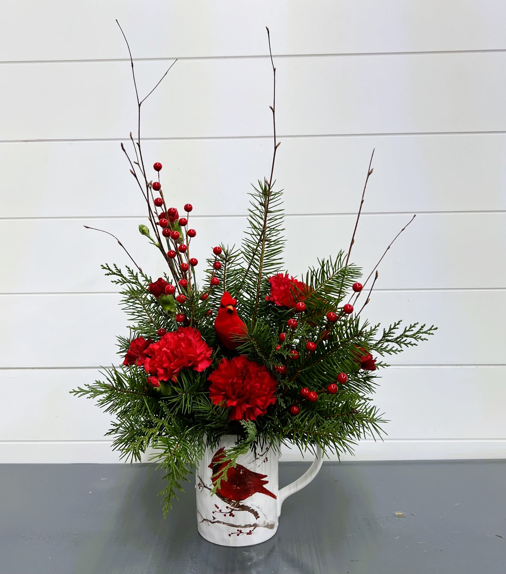 Cardinal In The Snow Mug Bouquet (LOCAL PICK UP AND DELIVERY ONLY) - Cardinal in the Snow Mug Celebrate the season with the Cardinal in the Snow Mug, a charming Christmas gift that combines festive blooms with seasonal cheer. This ceramic mug features bright red carnations, red hypericum berries, a striking red cardinal, and lush seasonal greenery—creating a beautiful holiday display perfect for gifting to friends, family, or co-workers.  Ideal as a thoughtful Christmas present or a festive centerpiece, this arrangement brings warmth and holiday joy to any home.  Note: Available for local pick-up and delivery only.
