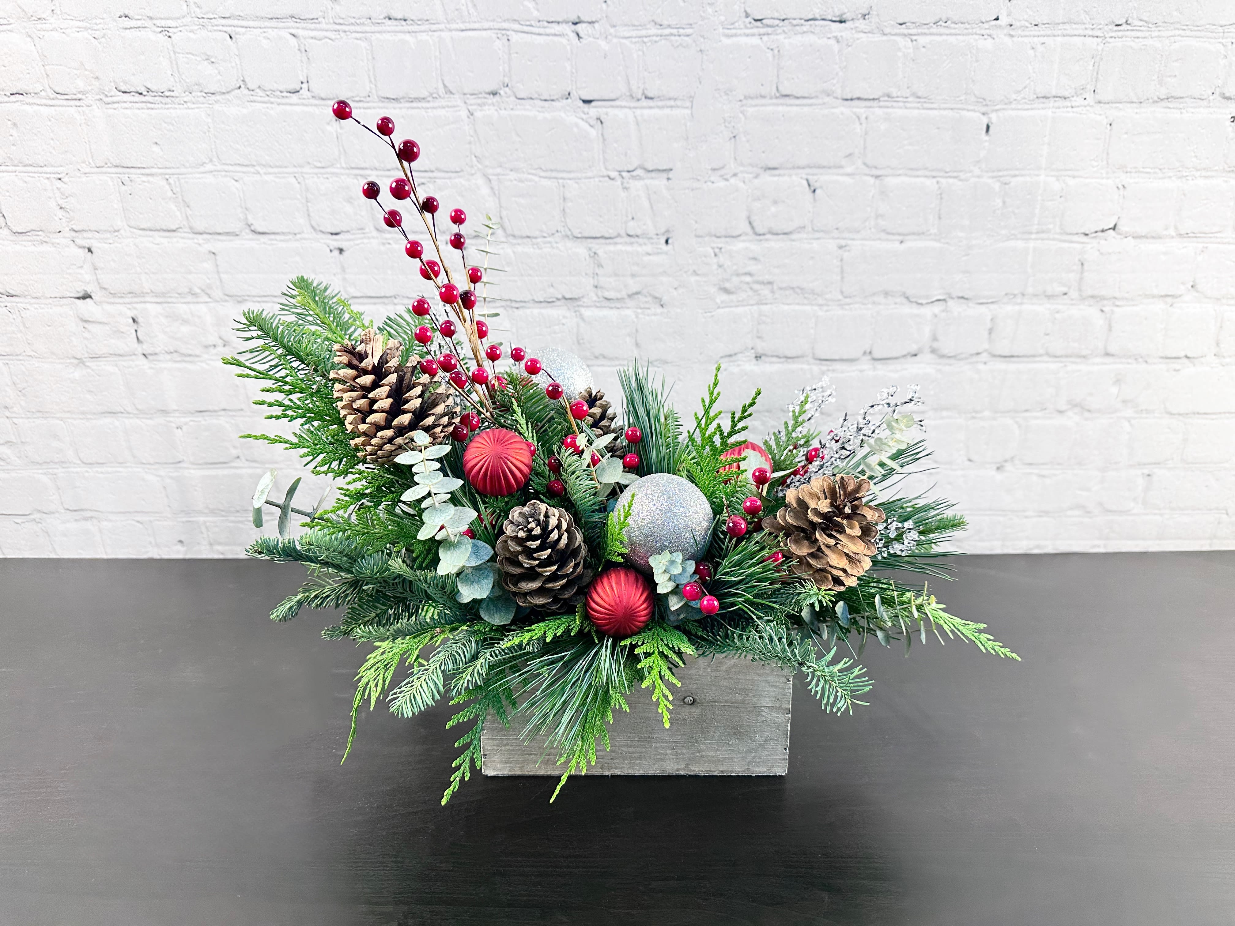 Yuletide Glow - Welcome in the scents of the season with this winter greenery arrangement! Consisting of various fragrant winter greenery, this arrangement will be sure to last through your holiday festivities with pinecones, ornaments, and berries adding in some pops of color and texture! This arrangement is available in standard and deluxe sizing. (NOTE: arrangement will be approximately 14"T x 18"L in a wood box container. Color of container and ornaments may vary from picture based on availability.)