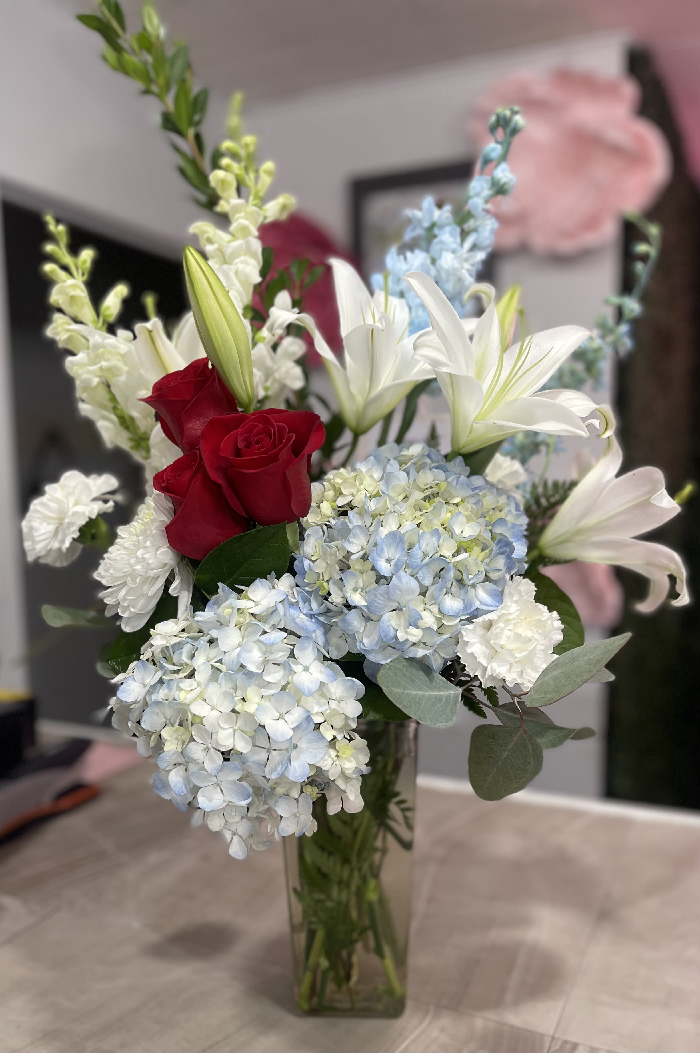 Stunning Stella - A collection of red roses, blooming white lilies, white carnations, white stock and blue delphinium come beautifully arranged in a clear vase.
