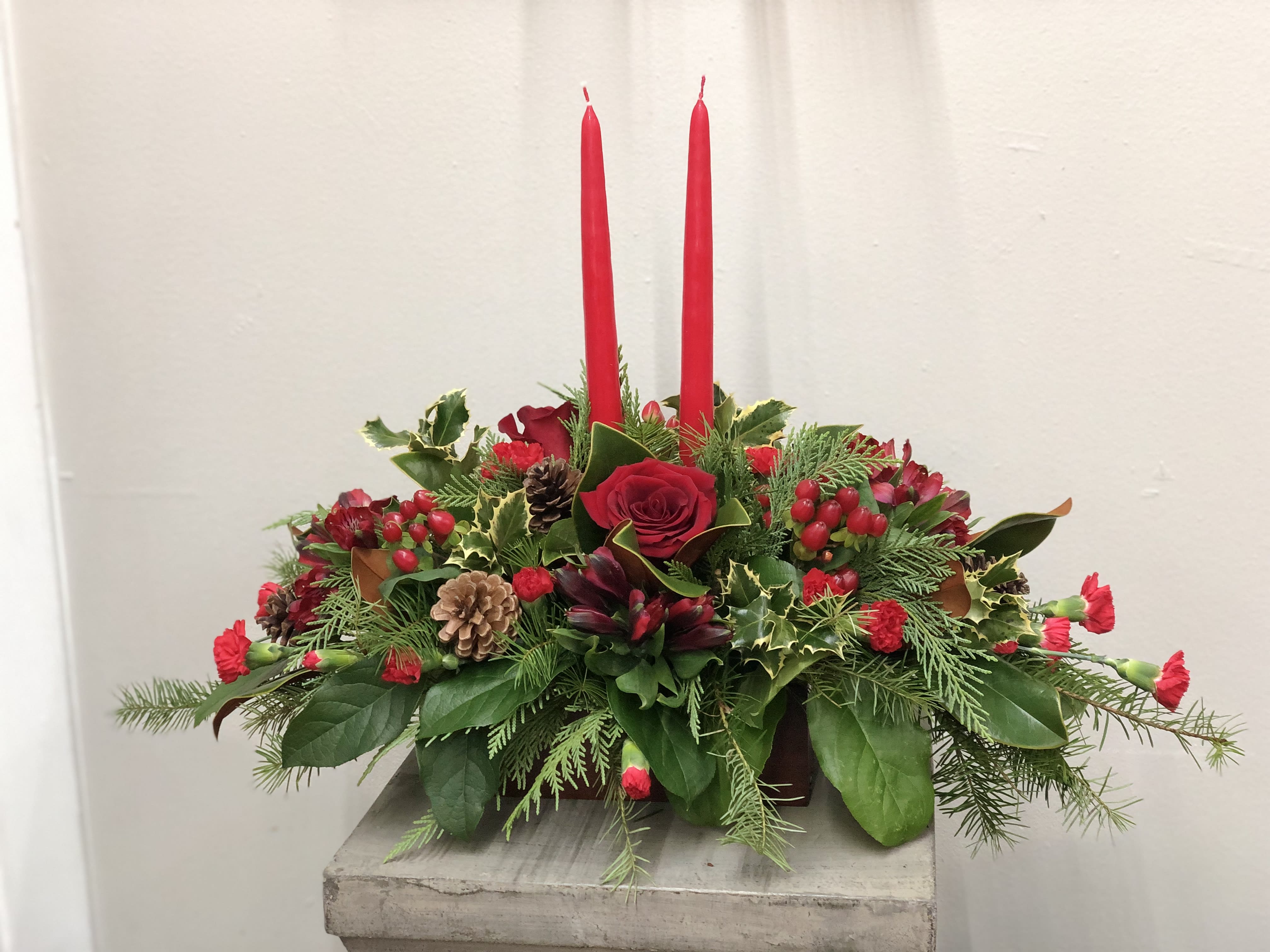 Seasons Greeting Centerpiece  - Seasons Greeting Centerpiece Adorn your holiday table with Teleflora’s Seasons Greeting Centerpiece, a festive low and long arrangement designed to delight. Featuring classic red roses, vibrant red carnations, seasonal red berries, and lush winter greens, this centerpiece is accented with elegant red candles for a warm, holiday glow. Perfect for holiday entertaining or gifting, it brings timeless seasonal charm to any celebration.