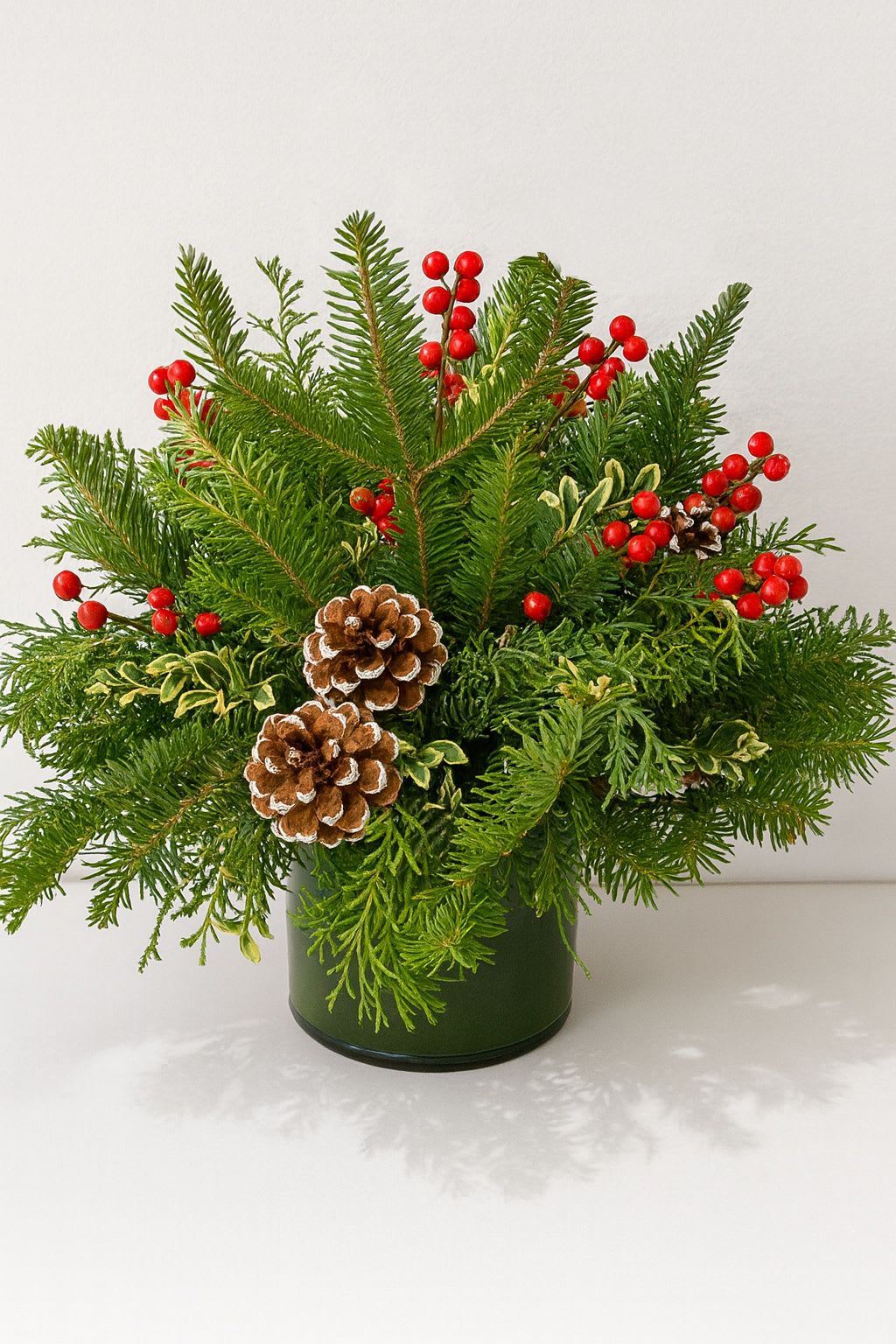 Woodland Berry &amp; Pine Pot - Bring the fresh feeling of a winter forest right into your home. The Woodland Berry &amp; Pine Pot is a celebration of nature’s winter textures. We’ve combined spiky spruce tips with soft, draping greens and touches of yellow-edged foliage to create a wild, freshly-gathered look. The star of the show is the contrast between the bright red berries and the snow-dusted pinecones, which add a touch of rustic charm to the display. Housed in a simple green container that lets the foliage shine, this arrangement is a cozy reminder of winter walks and holiday traditions. It’s compact enough for a desk but bold enough to brighten up a room. Perfect for: Coffee tables, home office desks, and &quot;thinking of you&quot; gifts.