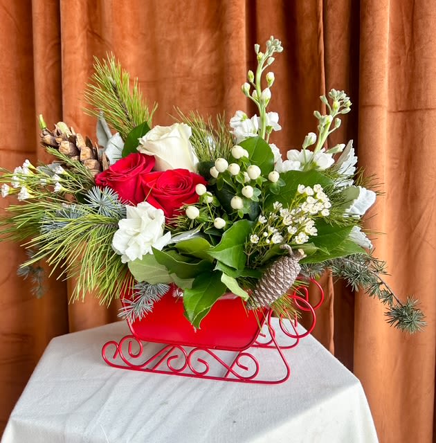 Santa's Sleigh - Celebrate the magic of the season with this charming winter centerpiece designed in a festive red metal sleigh. Lush evergreens and soft winter textures set the scene for vibrant red roses, creamy white blooms, and delicate accents of pinecones and snowberries. This arrangement brings the warmth and wonder of a classic holiday sleigh ride right to your tabletop. Perfect for holiday gatherings, gifting, or adding a timeless touch of Christmas cheer to your home. Container style may vary slightly based on availability, but the festive sleigh theme will remain.