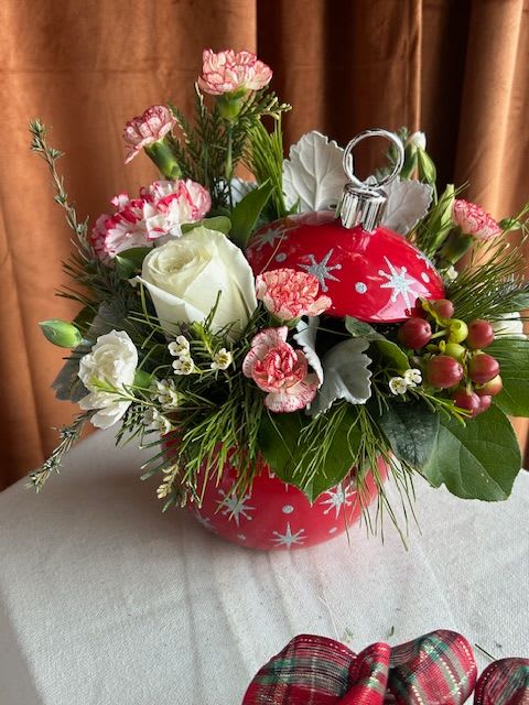 Holiday Cheer Ornament - Add a joyful pop of Christmas cheer to any space with this festive ornament-inspired floral design. Nestled inside a decorative holiday ornament container, this piece features a charming mix of winter greens, white roses, seasonal mini carnations, berries, and delicate textural accents. Fresh evergreens and soft blooms come together to create a playful yet elegant arrangement perfect for gifting, decorating desks and counters, or bringing a touch of magic to your holiday celebrations.
