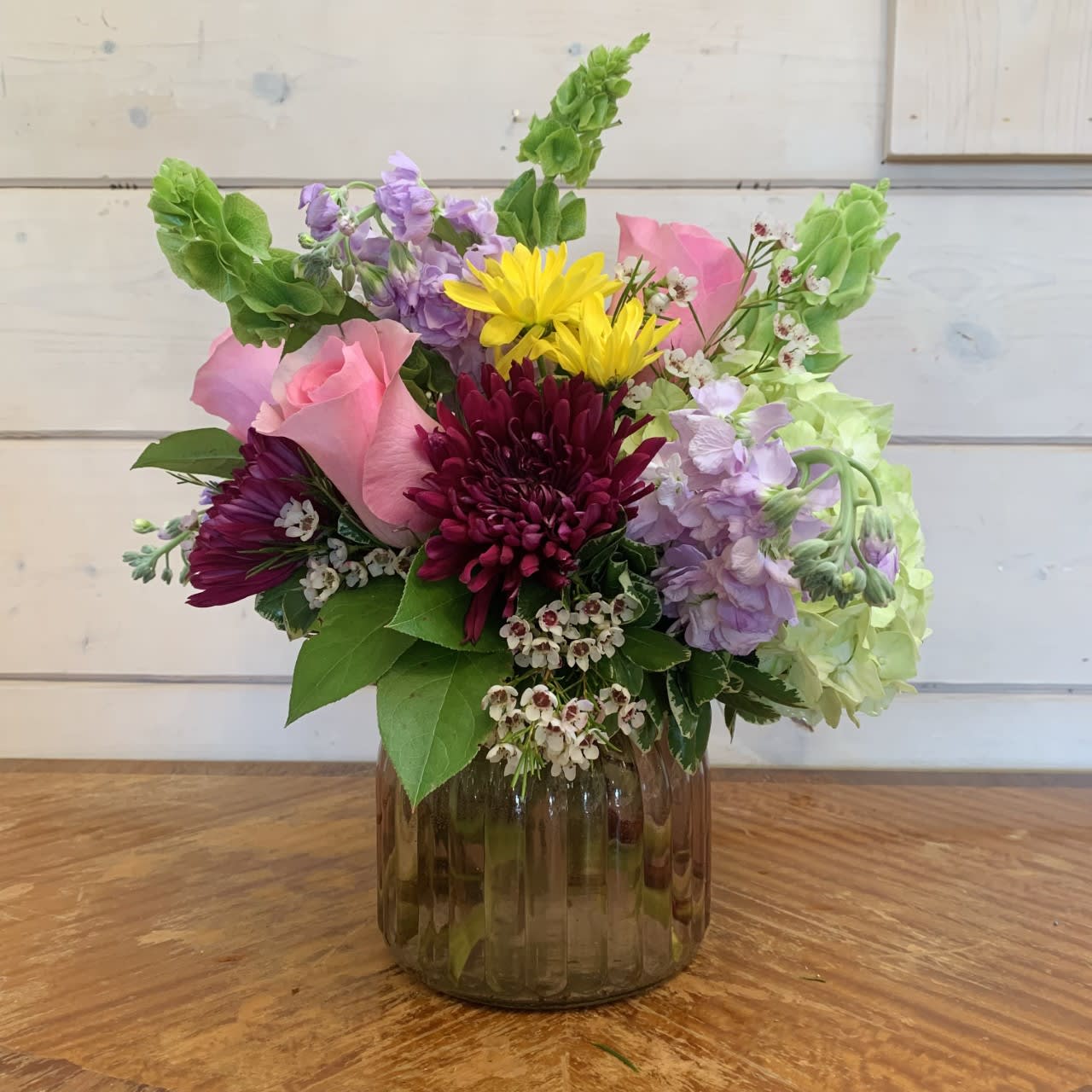 A Touch of Spring  - Green hydrangea, bells of Ireland, lavender stock, pink roses, purple cremon chrysanthemums and yellow daisies are arranged in an extra large pink ginger jar with white wax flower.  Approximately 14in tall and 12in wide.  