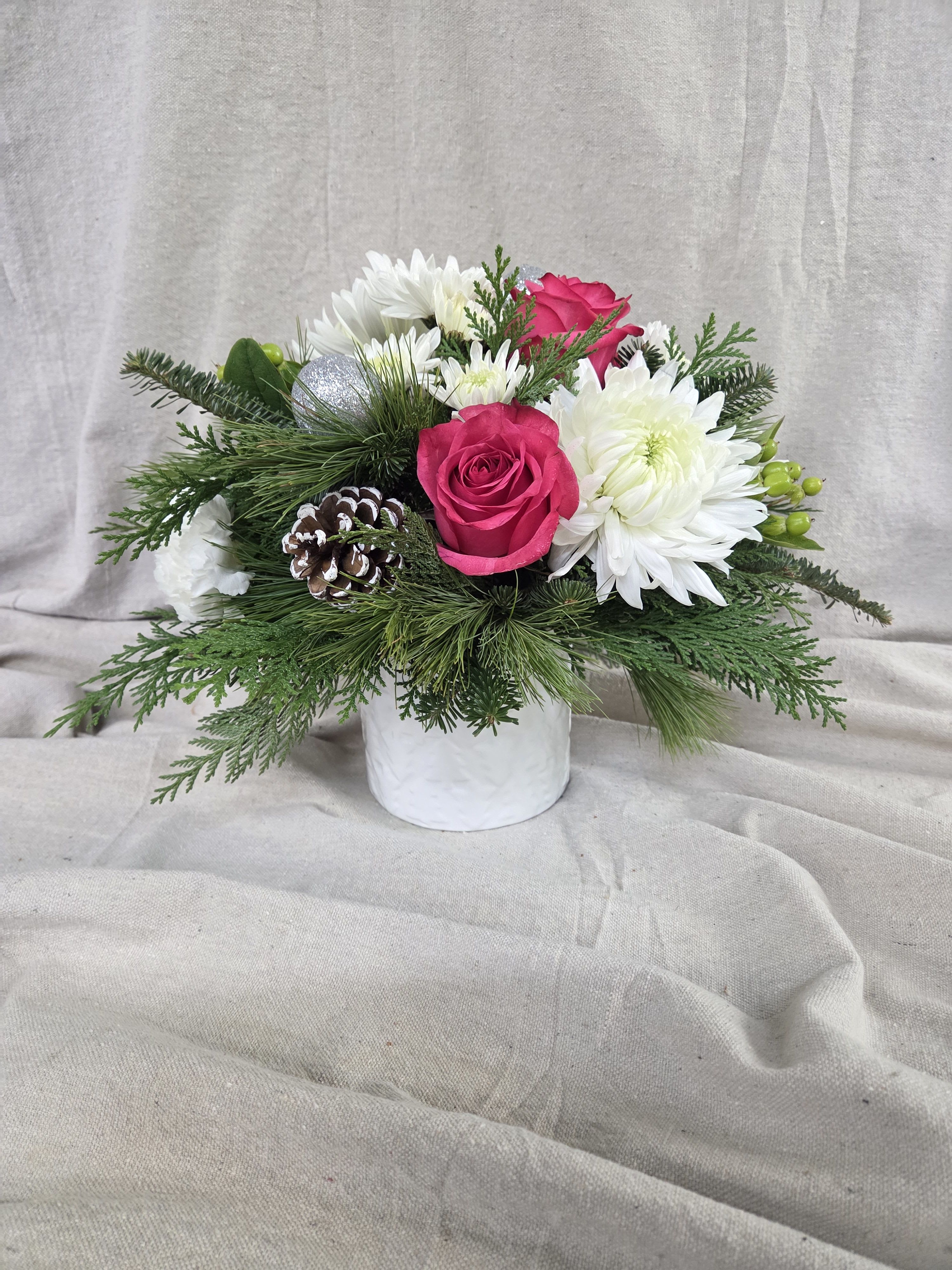 Blushing Winter Arrangement  - Add a pop of holiday cheer with this festive Christmas arrangement. Pink and white blooms are paired with lush winter greens, creating a vibrant and seasonal display. Arranged in a white vase decorated with holiday trees, this centerpiece brings color, joy, and classic Christmas charm to any table, mantel, or gift-giving occasion.