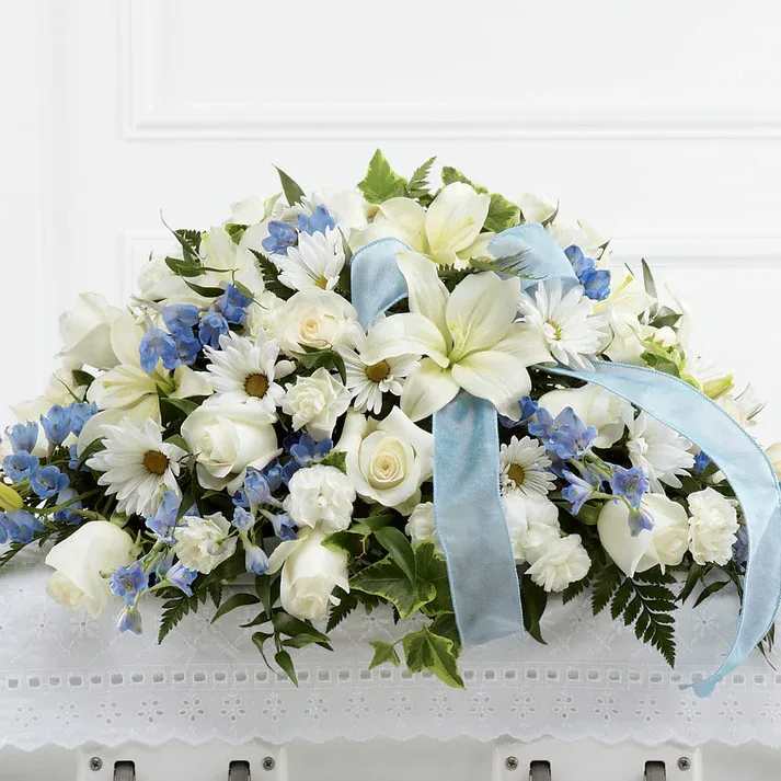 Tender Treasure Casket Spray - The Tender Treasure Casket Spray is a beautiful way to honor the life of the child taken too soon. White roses, daisies, monte casino asters, Asiatic lilies, and mini carnations are accented with light blue delphinium and a light blue organza ribbon to be displayed on top of the casket at their final farewell service.  Details 15"h x 25"w
