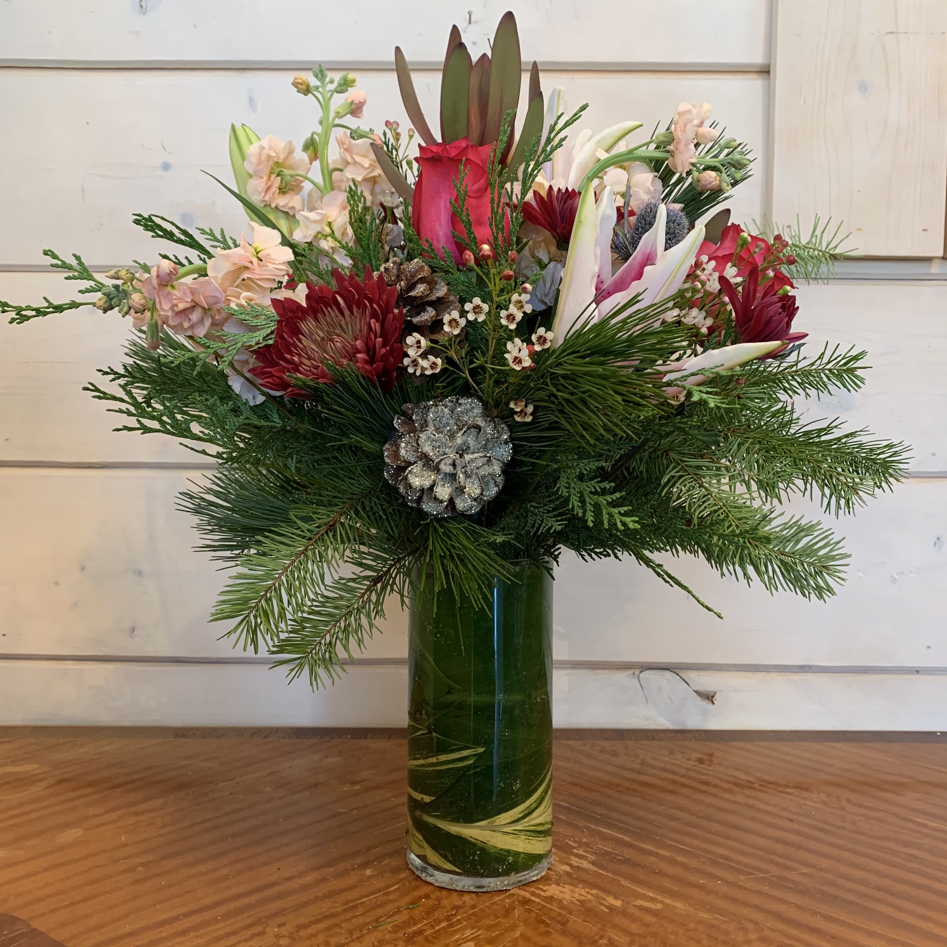 Love and Laughter  - Hot pink roses, burgundy chrysanthemums, peach stock, blue thistle, and stargazer lilies arranged in a 9in leaf lined cylinder with seasonal evergreens, white wax flower and glitter pinecones. 