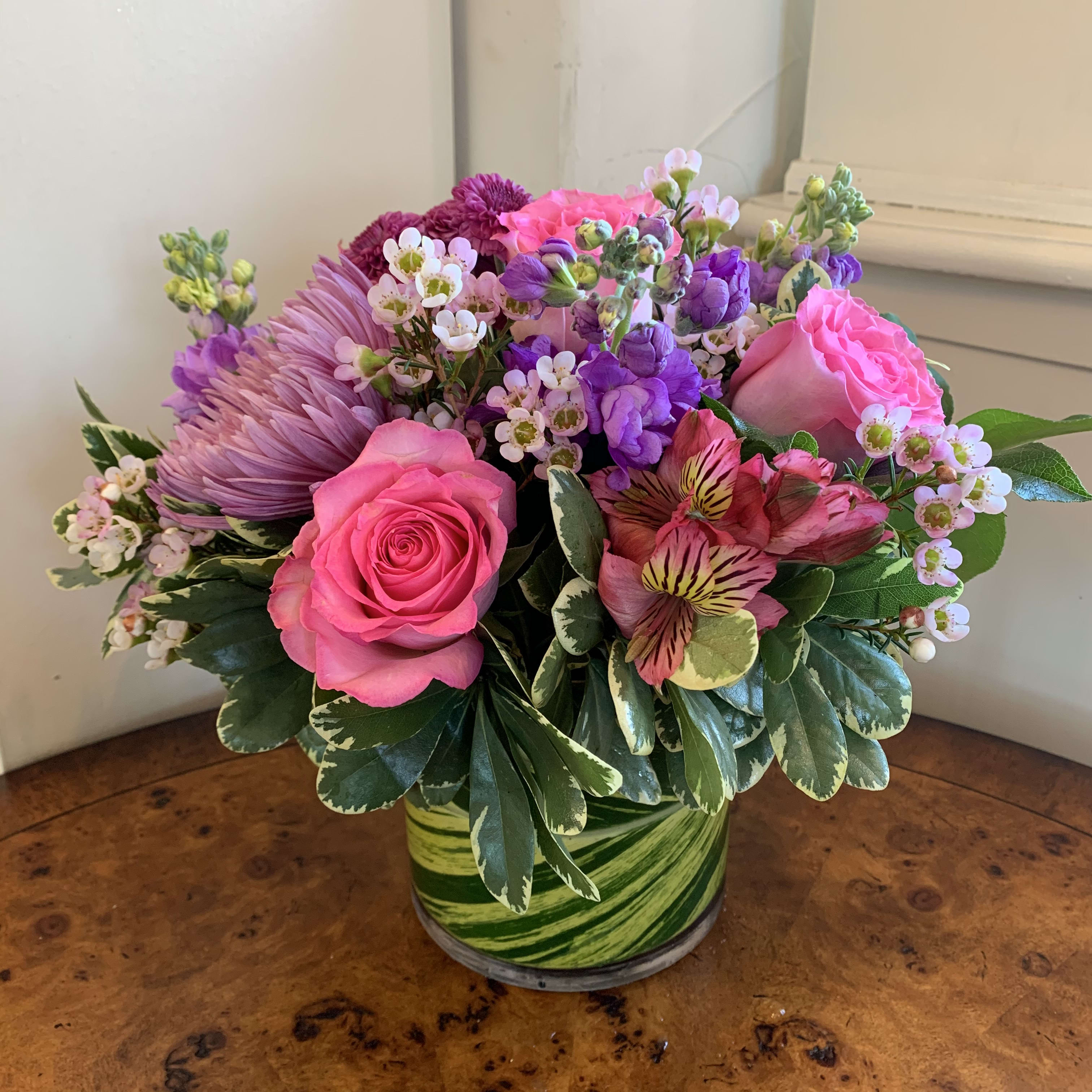 Head Over Heels  - Pink roses, medium purple stock, purple pom buttons, hot pink alstromeria, and lavender spider mum arranged with pink waxflower in a leaf lined small gather vase. Approximatly 10" tall by 8" wide. 