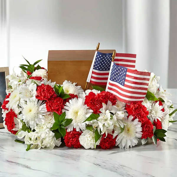 Spirit of Patriotism Cremation Adornment - Honor the life of a hero with a tribute in the colors and designs that reflect the nation they so proudly served. This stunning urn adornment sits table top with cremon mums, carnations and alstroemeria to hold an urn or tribute in the center. Our talented florists craft each bloom together and add two American flags to accent the patriotic blooms.  Details 10"h x 20"w Urn shown is not included.